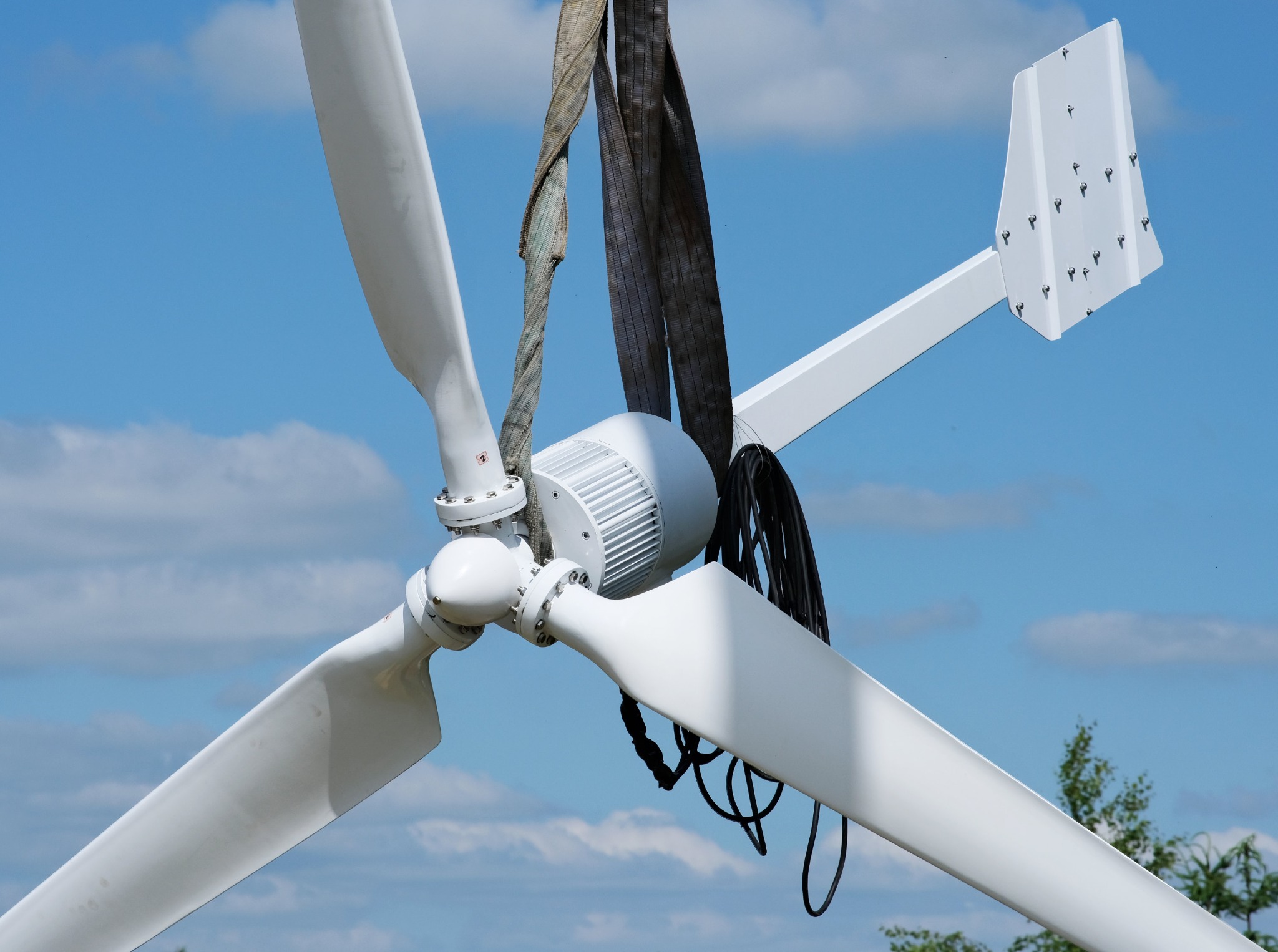 Biała turbina wiatrowa z trzema łopatami i statecznikiem pionowym, podnoszona za pomocą pasów transportowych na tle błękitnego nieba z chmurami.