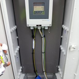 ASVOLT USŁUGI ELEKTRYCZNE ADRIAN SZCZĘCH - Otwarta szafka elektryczna z zamontowanym urządzeniem pomiarowym Endress+Hauser Promag, widoczne kable w peszlu i z etykietą informacyjną, ręka w folii aluminiowej w lewym dolnym rogu.