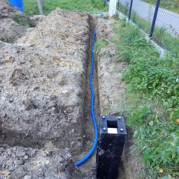 ASVOLT USŁUGI ELEKTRYCZNE ADRIAN SZCZĘCH - Wykop w ziemi z ułożoną niebieską rurą karbowaną, czarna skrzynka z otworami na górze, widoczny fragment ogrodzenia panelowego i trawy.