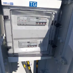Otwarta skrzynka elektryczna z przezroczystą pokrywą, widoczne bezpieczniki Siemens, oznaczenia 'WYL. GŁÓWNY' i 'TABLICA GNIAZD', kable w peszlach z żółtymi oznaczeniami u dołu skrzynki, napis 'TG'...