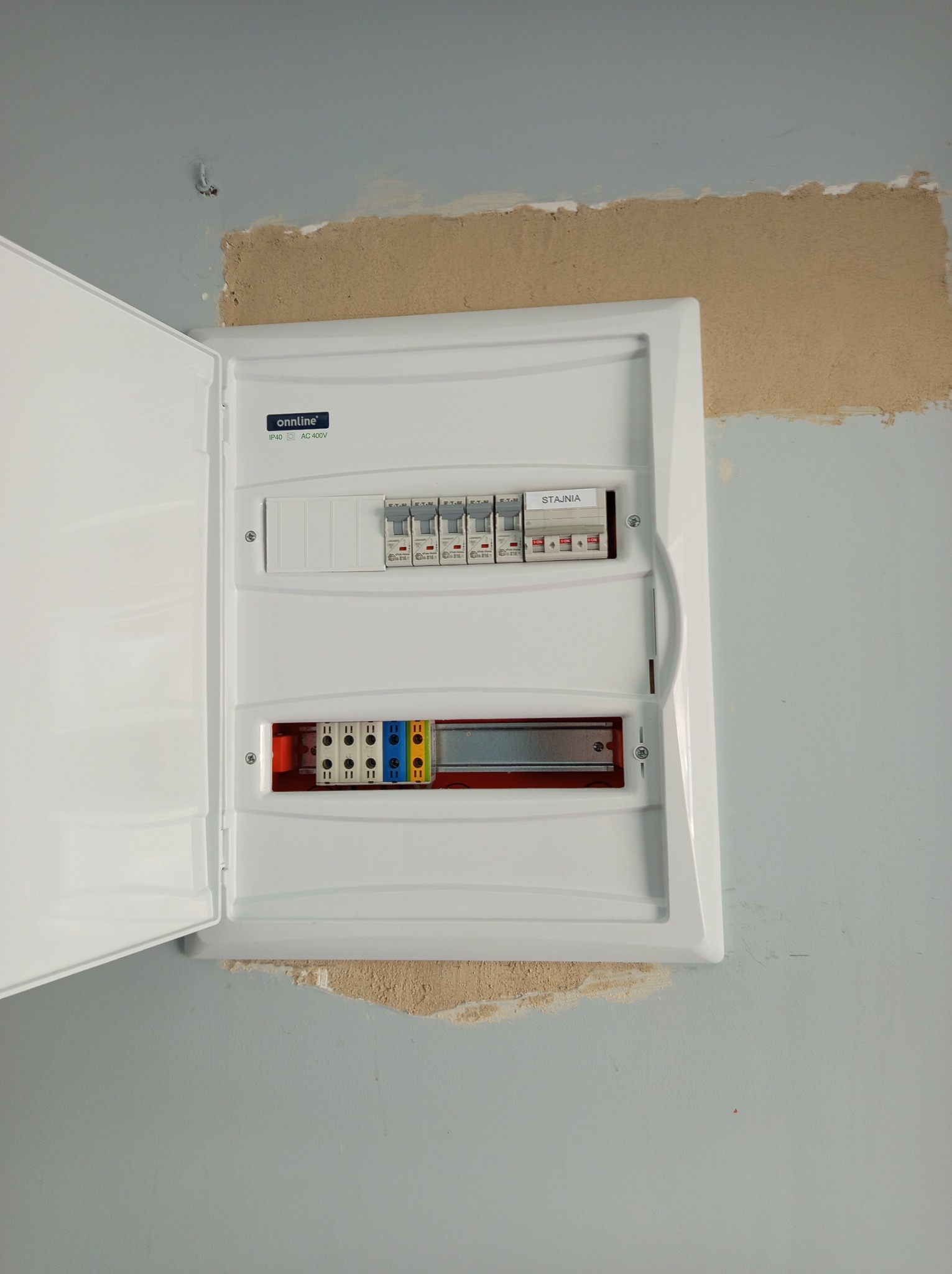 Otwarta, biała skrzynka rozdzielcza z bezpiecznikami i listwami zaciskowymi, zamontowana na nieestetycznej, szarej ścianie z widocznymi śladami po naprawach.