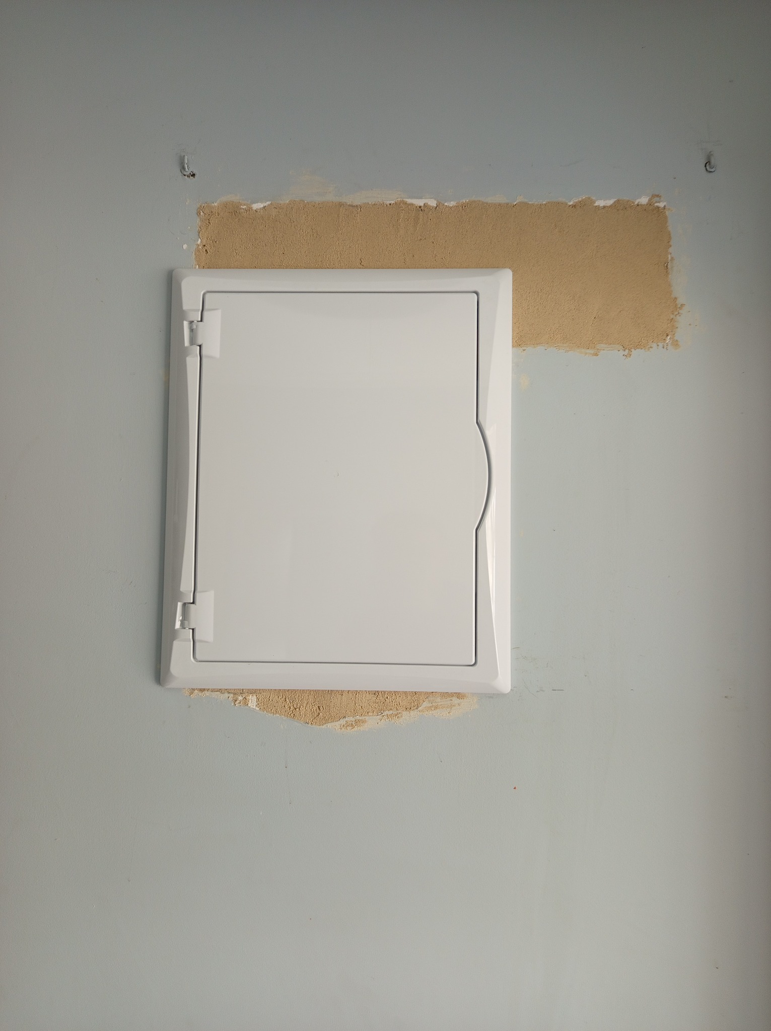 Biała klapka skrzynki elektrycznej na szarej ścianie z ubytkami tynku wokół, widoczne ślady po wierceniu.