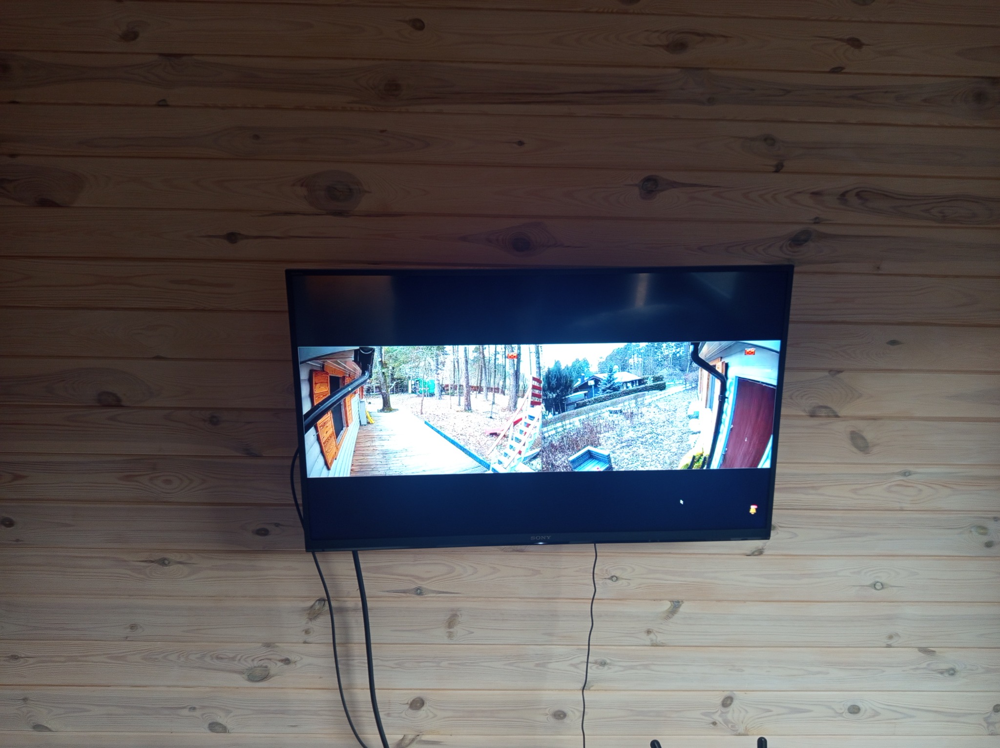 Ekran telewizora LCD zamontowanego na ścianie z boazerii, wyświetlający obraz z kamery monitoringu obejmującej podwórko z domem, drzewami i ścieżką.