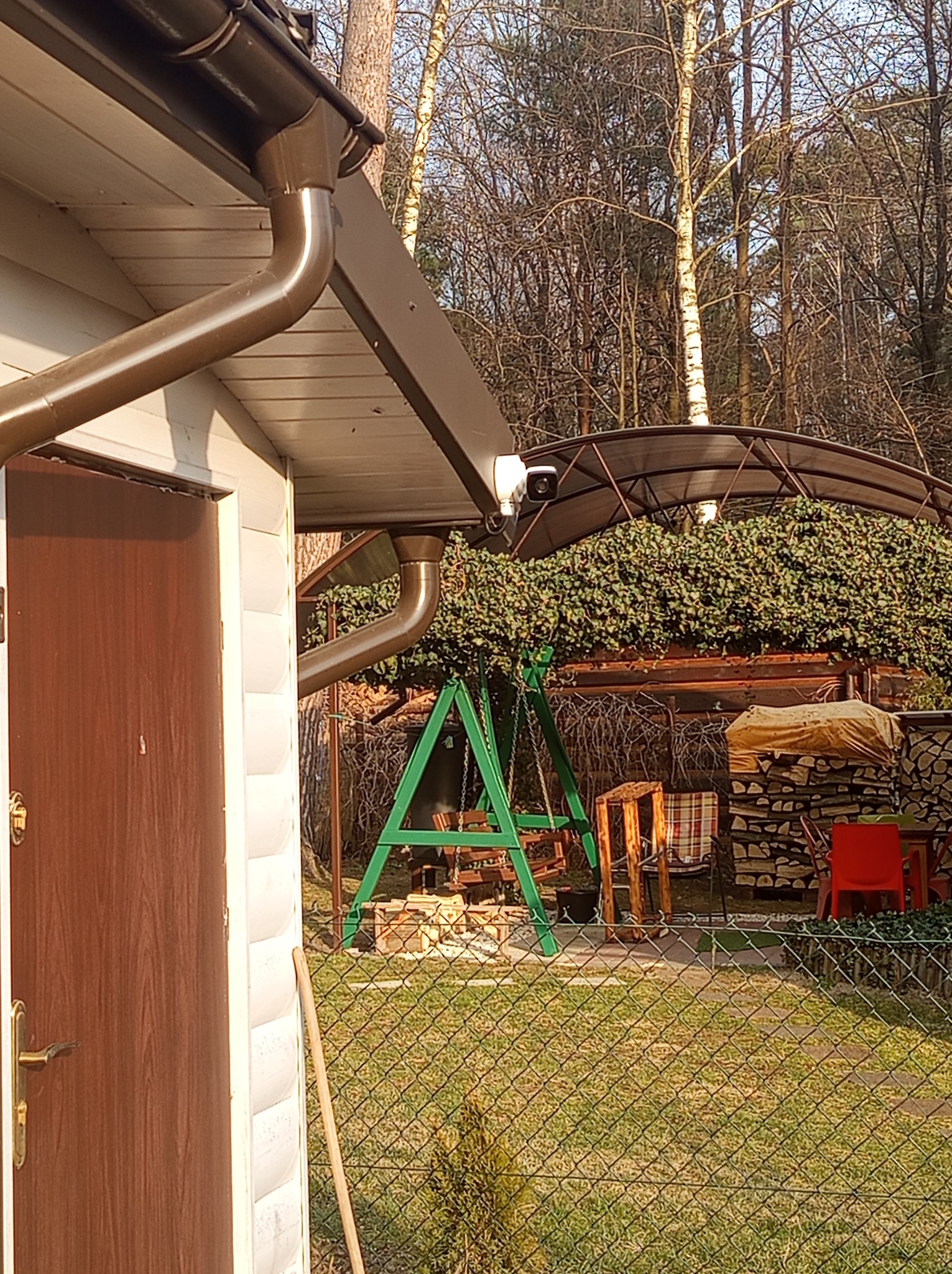 Kamera monitoringu zamontowana pod brązowym orynnowaniem budynku gospodarczego z widokiem na podwórko z huśtawką i drewutnią.