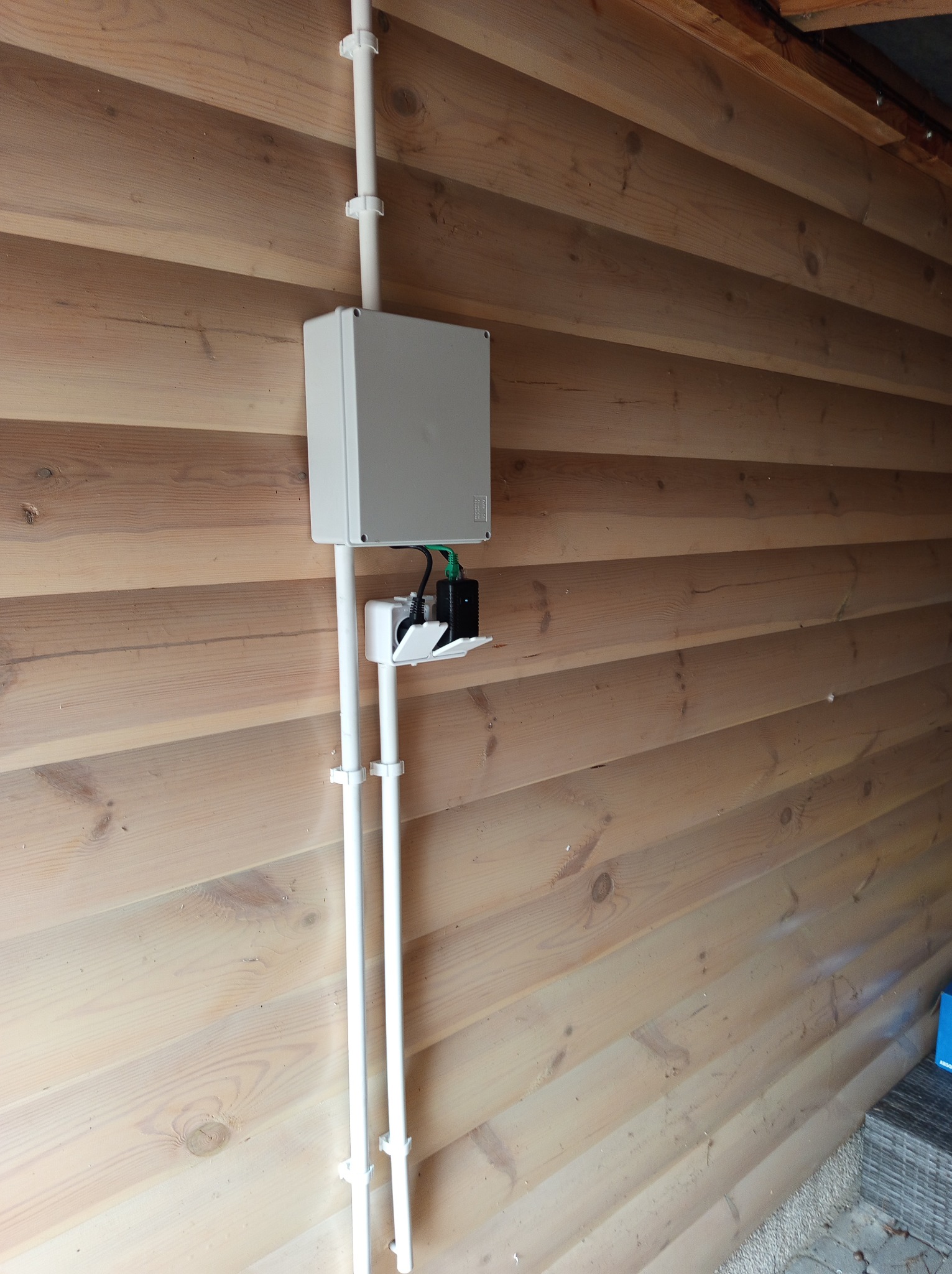 Szara skrzynka elektryczna z białymi rurkami PCV na drewnianej ścianie z widocznym gniazdem i wtyczką pod spodem.