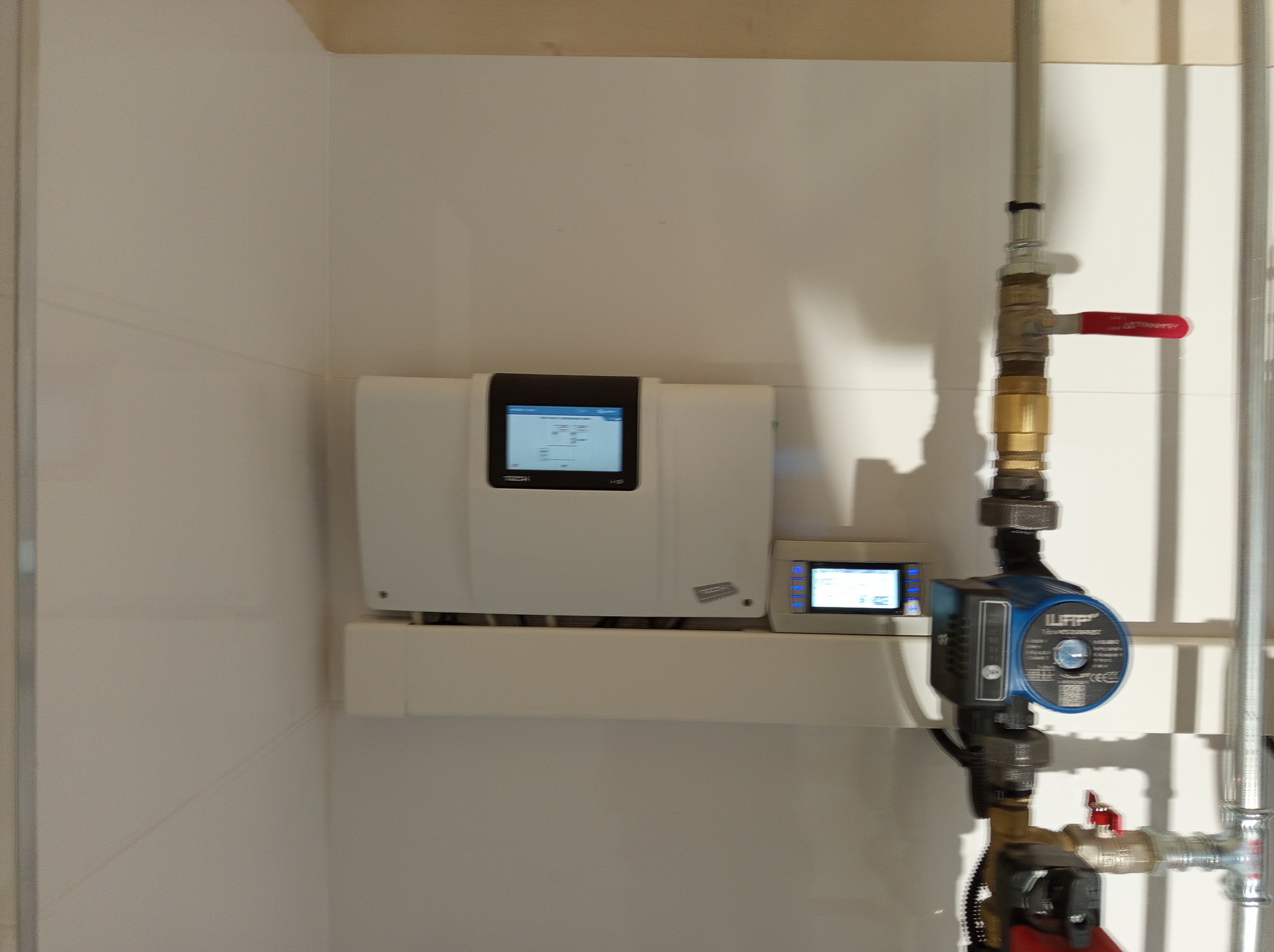 Biała skrzynka sterownicza z czarnym ekranem i niebieskim wyświetlaczem, zamontowana obok pompy obiegowej z zaworami i rurami, na tle białej ściany.