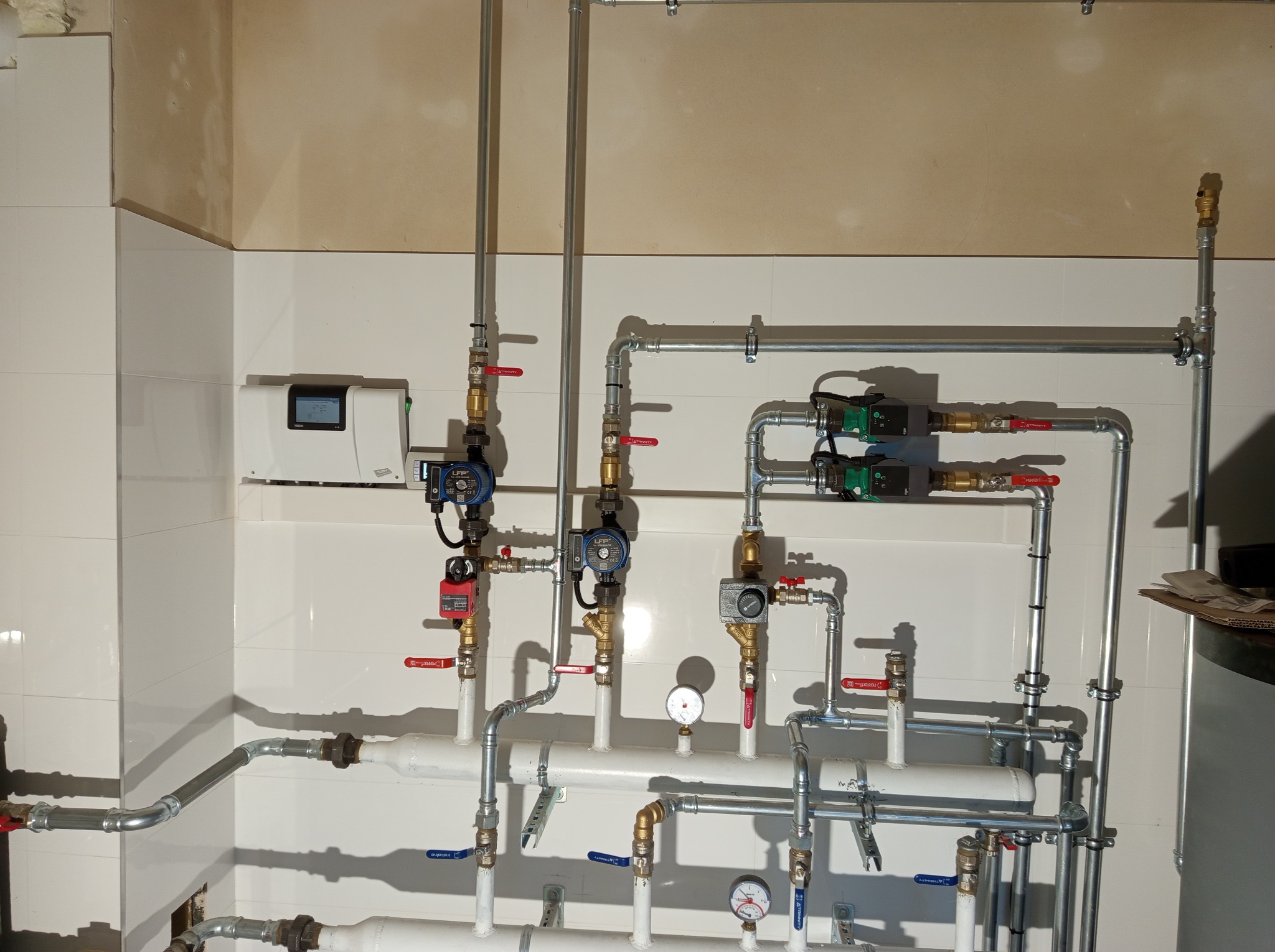 Rozbudowana instalacja centralnego ogrzewania z licznymi rurami, zaworami, pompami obiegowymi i manometrami zamontowana na ścianie pokrytej białymi płytkami w pomieszczeniu w trakcie wykańczania.