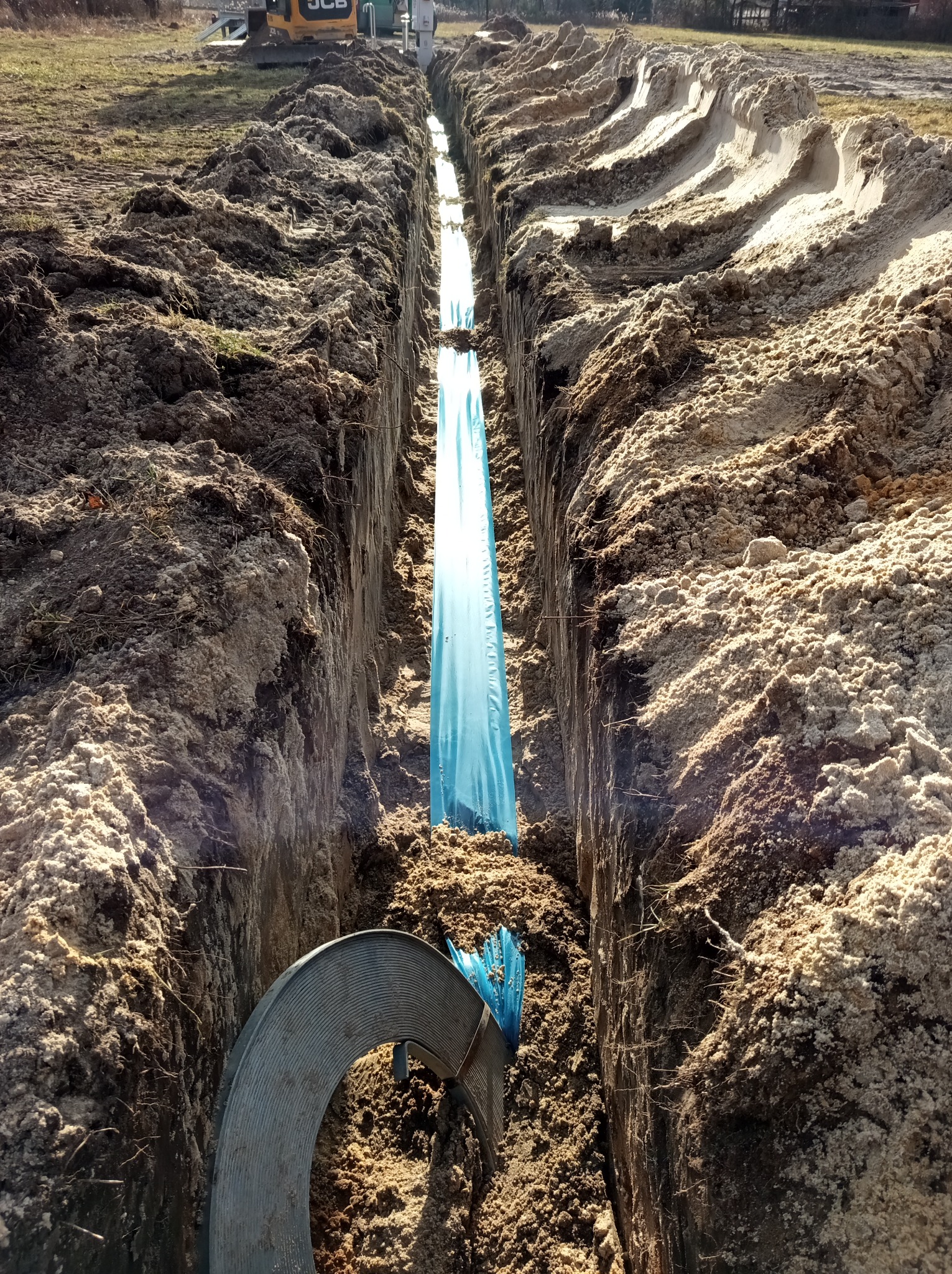 Wykop w ziemi z niebieską folią ochronną i fragmentem rury karbowanej na dnie, w tle widoczna koparka.