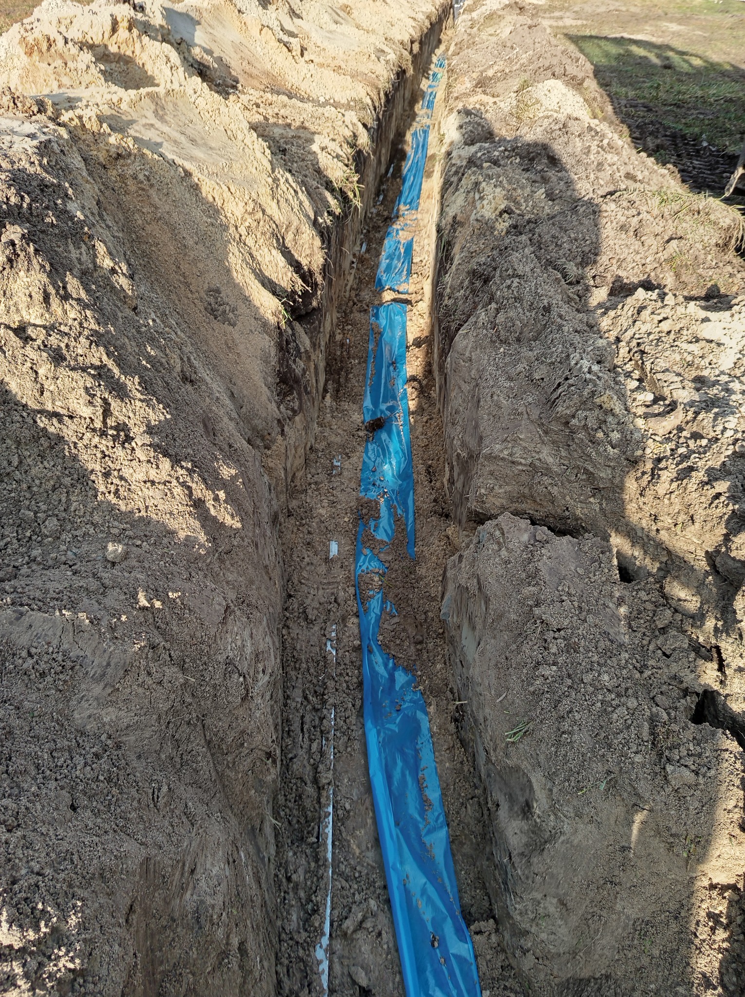 Wykop w ziemi wyłożony niebieską folią, prawdopodobnie przygotowany pod kable lub rury.