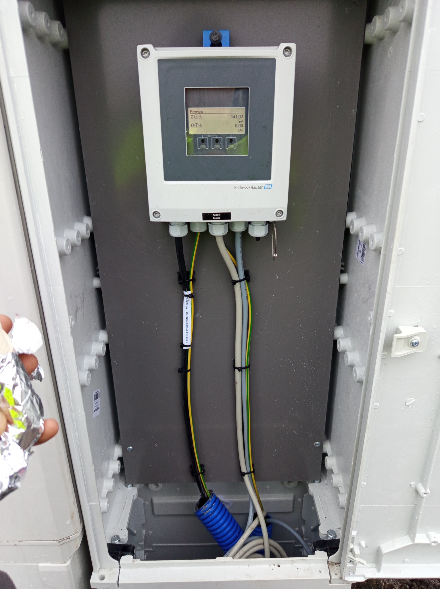 Otwarta szafka elektryczna z zamontowanym urządzeniem pomiarowym Endress+Hauser Promag, widoczne kable w peszlu i z etykietą informacyjną, ręka w folii aluminiowej w lewym dolnym rogu.