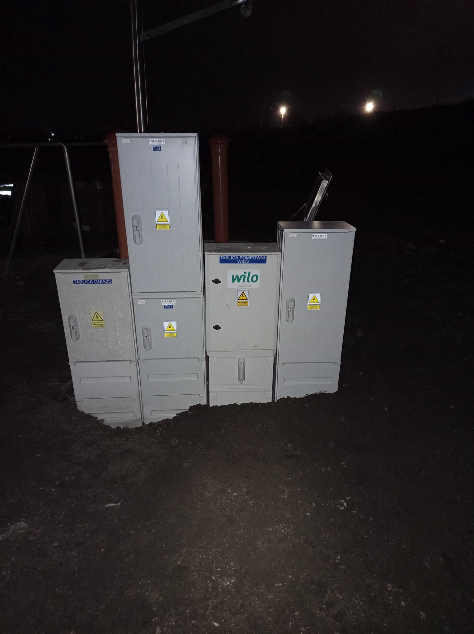 Zestaw szarych skrzynek elektrycznych różnych rozmiarów, ustawionych na ziemi w ciemności, z widocznymi tabliczkami ostrzegawczymi o napięciu elektrycznym i oznaczeniami producentów, oświetlonych...