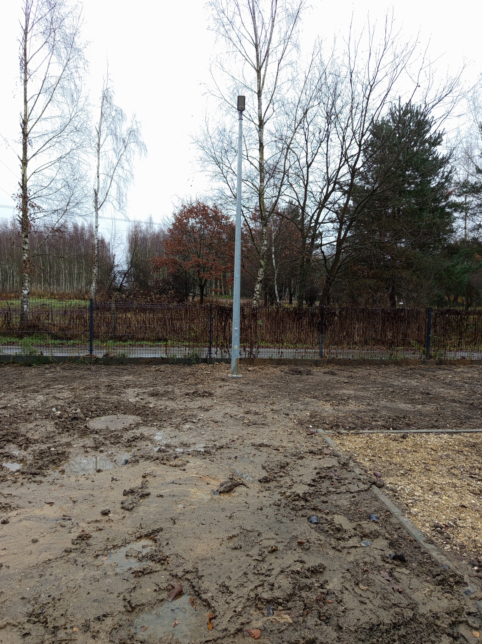 Metalowy słup oświetleniowy na terenie budowy z błotnistym podłożem i widocznym ogrodzeniem w tle, drzewa bez liści i pochmurne niebo.
