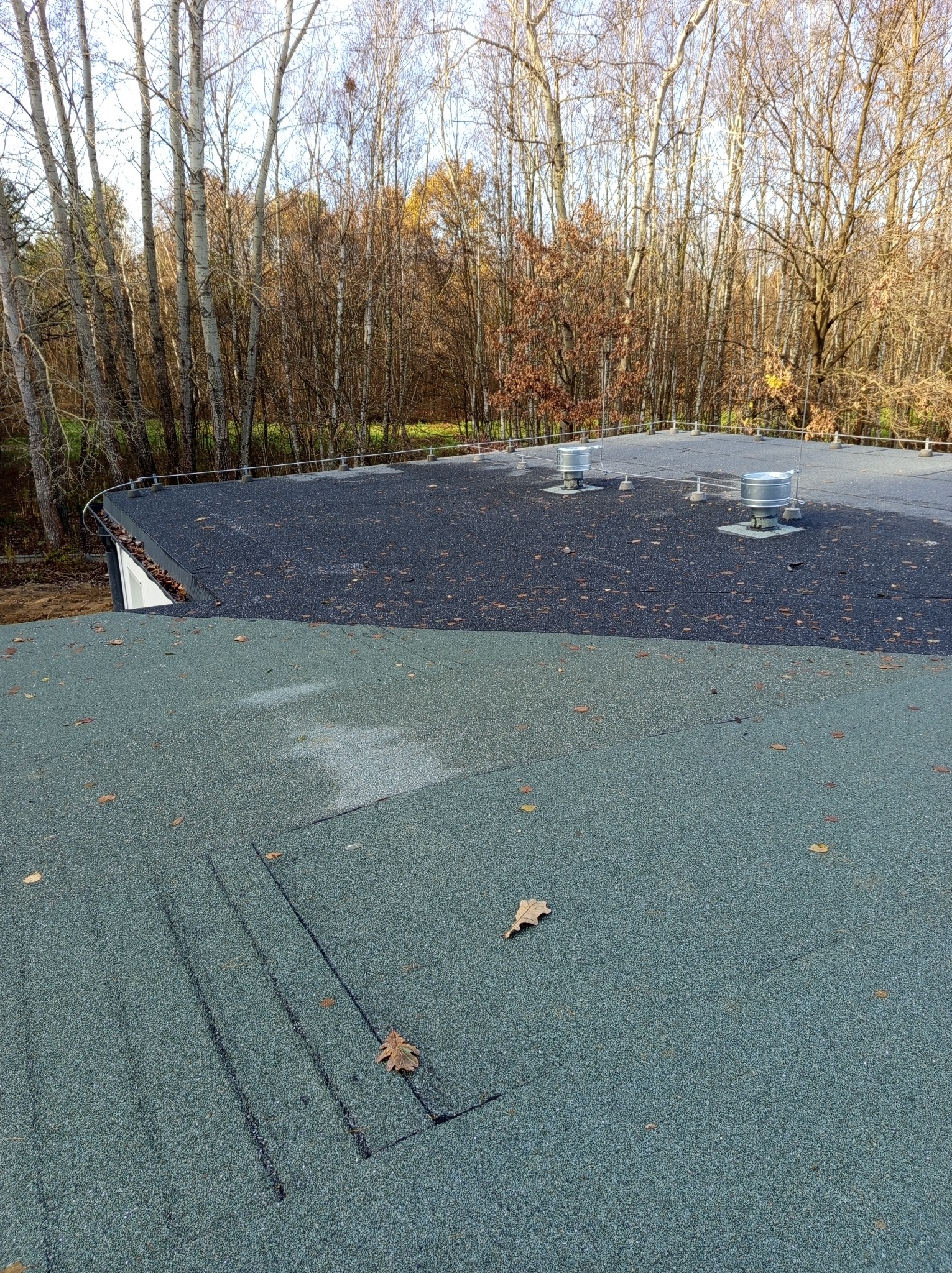 Widok z góry na płaski dach pokryty dwoma rodzajami papy: ciemnoszarą i zielono-szarą, z widocznymi odpowietrznikami i systemem odgromowym, w tle drzewa bez liści.