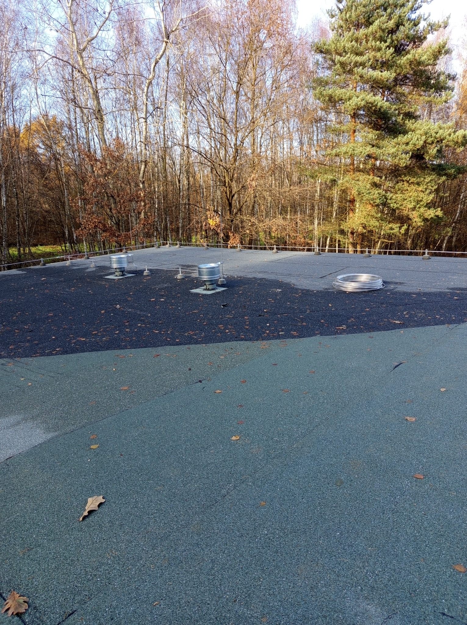 Płaski dach pokryty papą z widocznymi elementami instalacji odgromowej, takimi jak metalowe maszty i przewody, na tle jesiennego lasu.