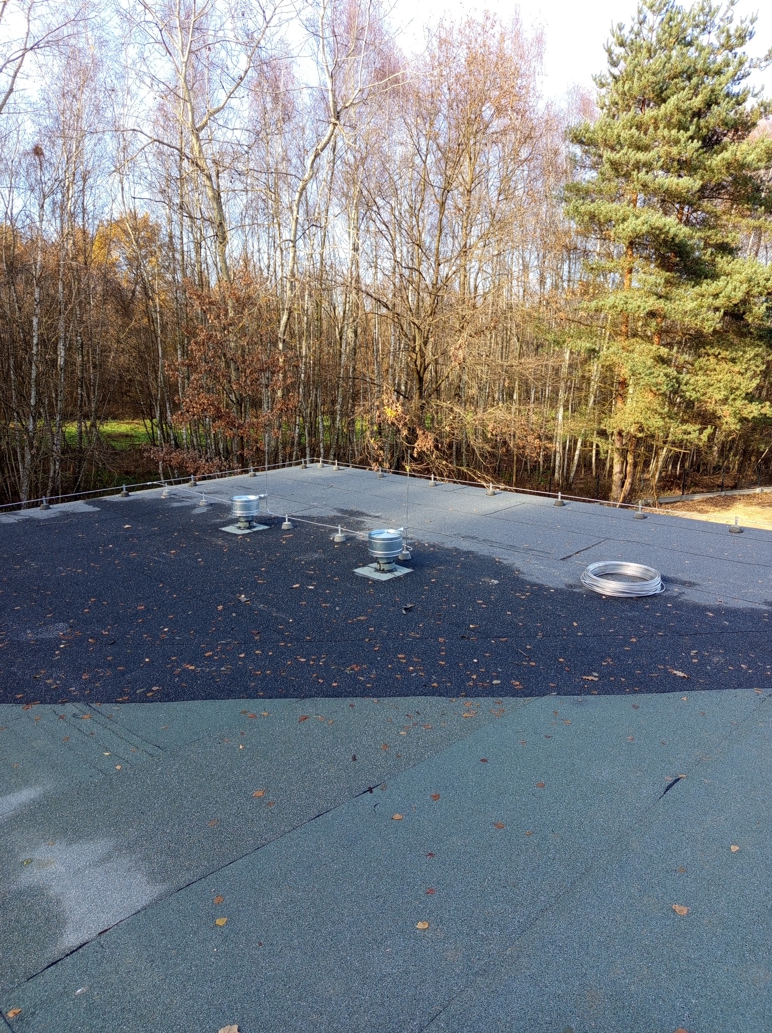 Płaski dach pokryty papą z elementami instalacji odgromowej: zwody pionowe z metalowymi osłonami, przewody odprowadzające i rolka zwiniętego drutu. W tle drzewa bez liści i iglaste.