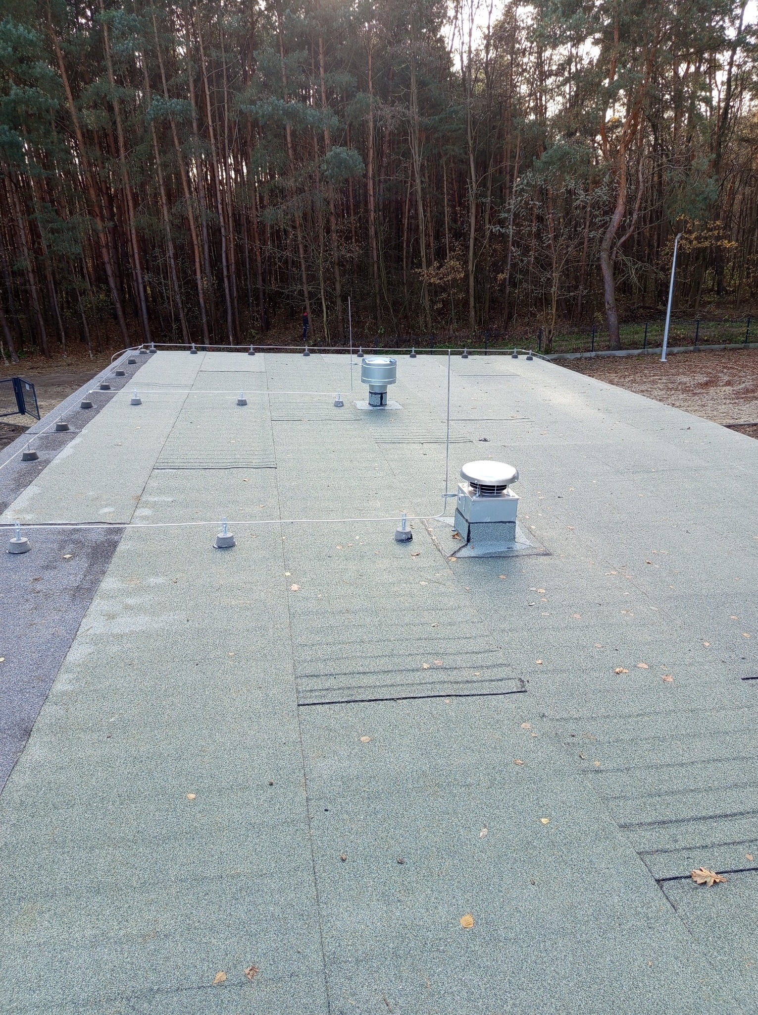 Płaski dach pokryty papą z widocznymi elementami instalacji odgromowej: uziomy i metalowe kominki wentylacyjne na tle lasu.