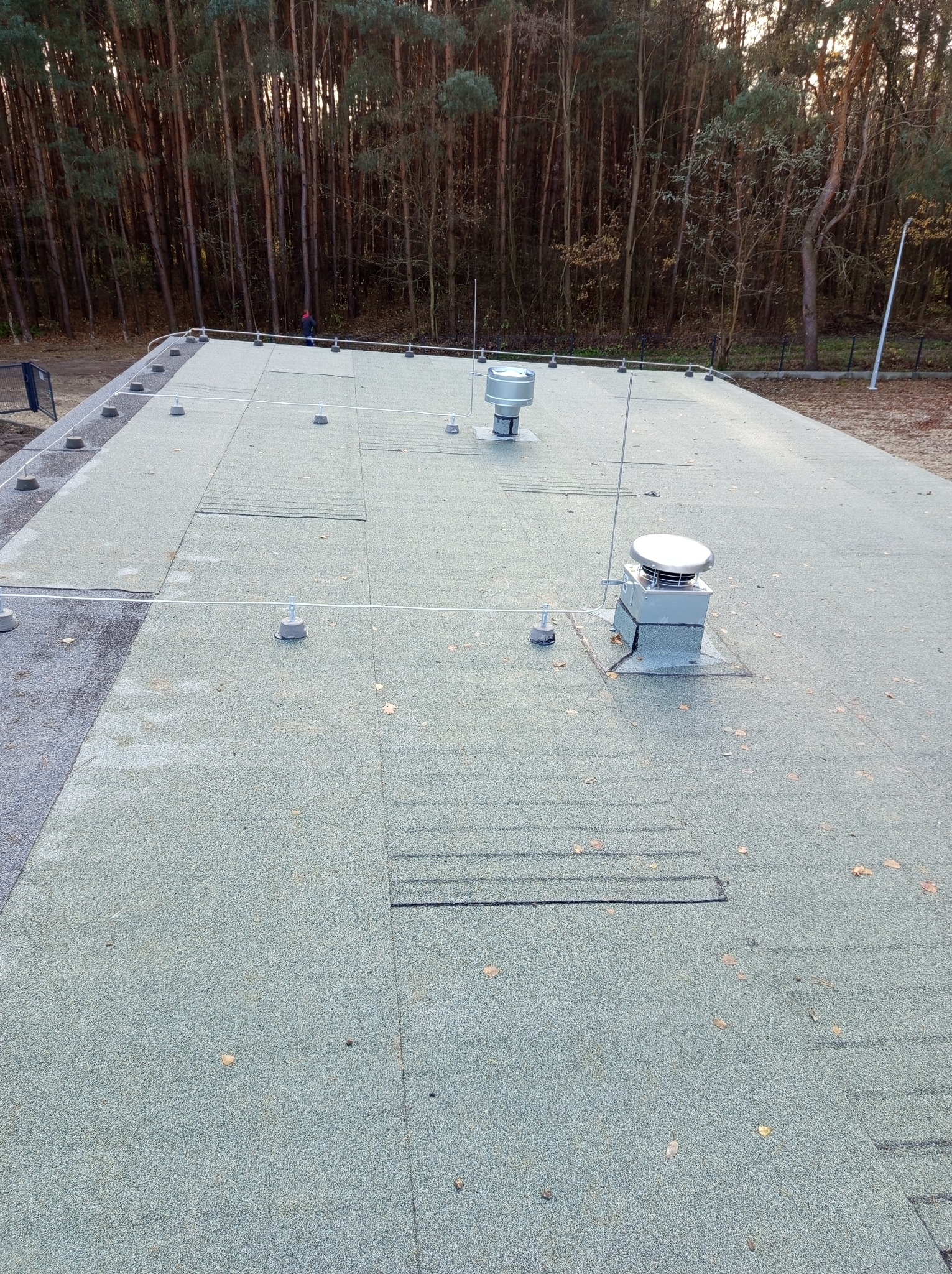 Płaski dach pokryty papą z zamontowanymi odgromnikami i wentylatorami, na tle lasu.