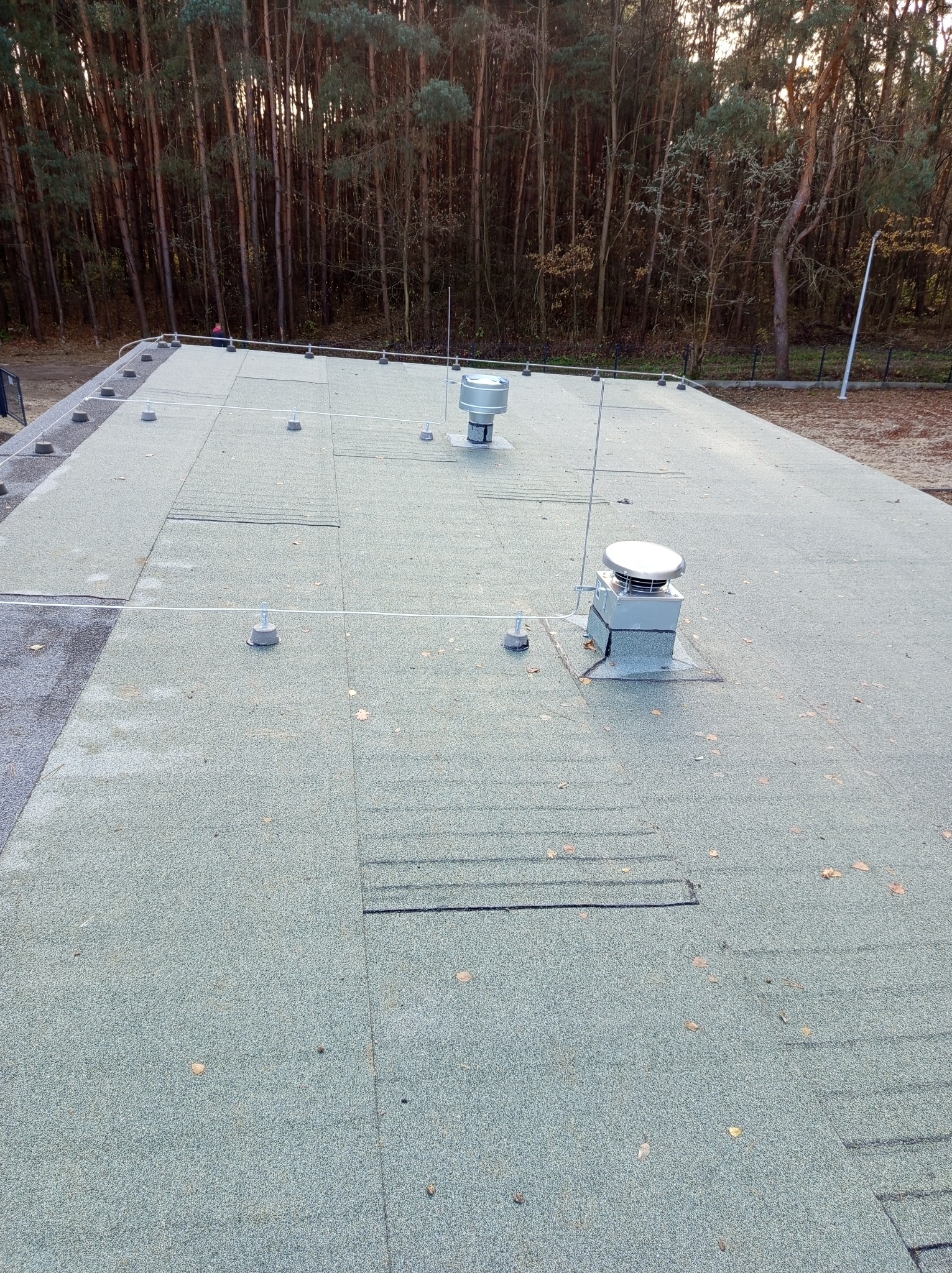 Płaski dach budynku pokryty papą z zainstalowaną instalacją odgromową, widoczne zwody pionowe i przewody odprowadzające, tło stanowią drzewa.