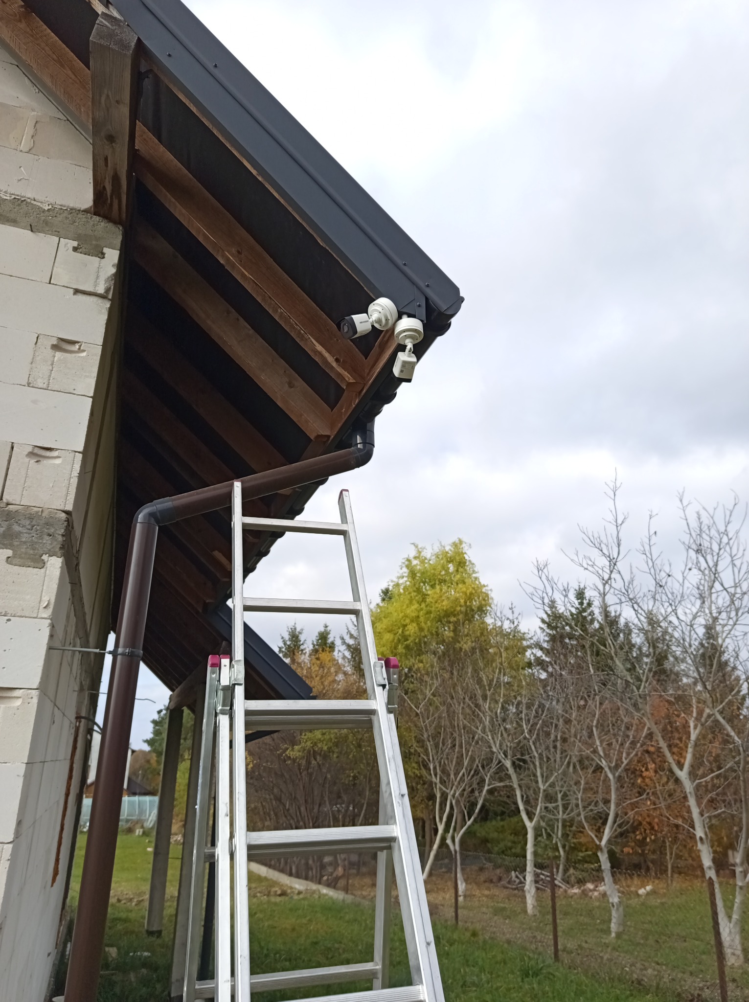Dwie białe kamery monitoringu zamontowane pod okapem dachu, widoczne z poziomu drabiny opierającej się o ścianę budynku w trakcie instalacji.