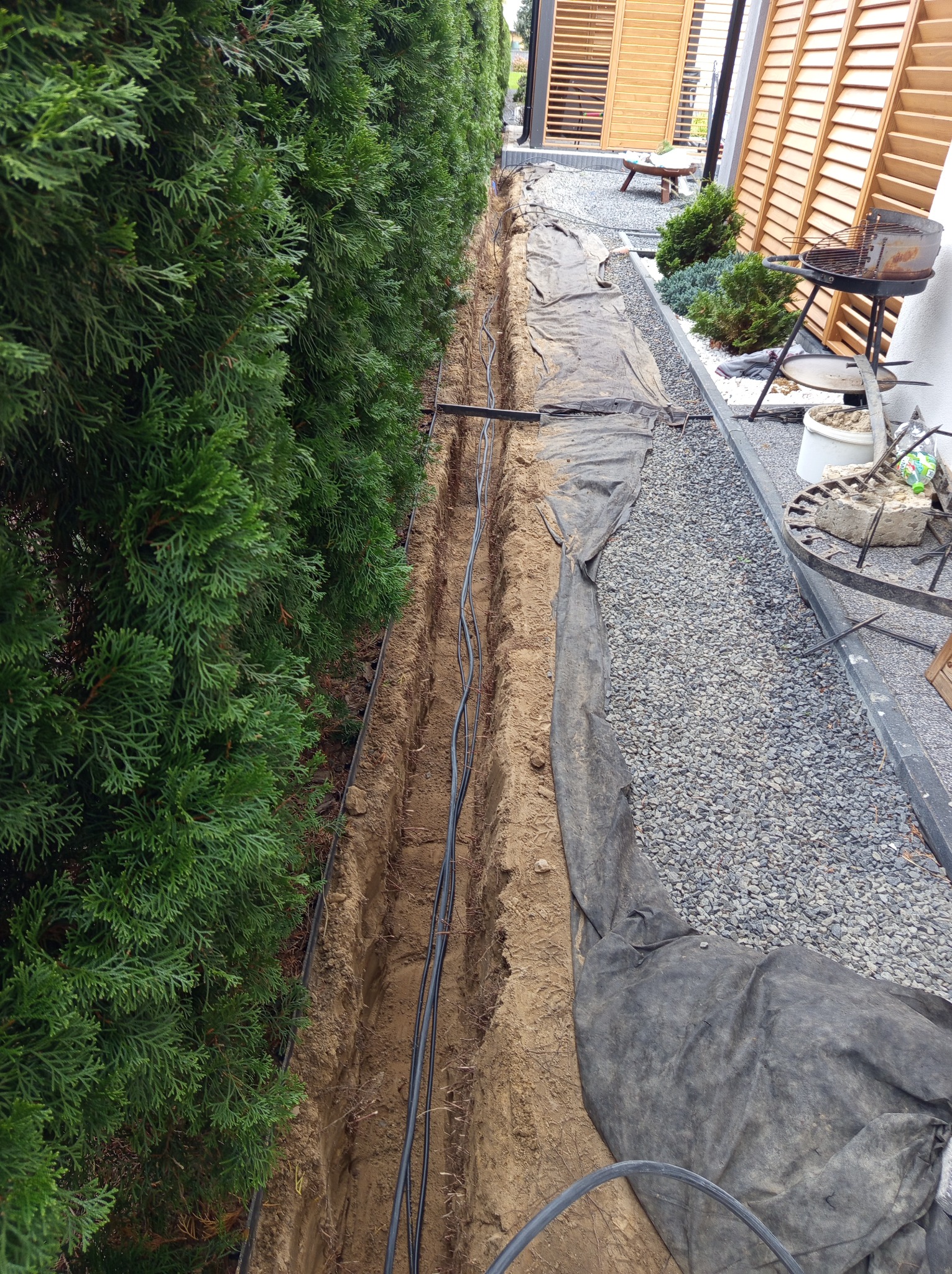 Wykop wzdłuż żywopłotu z ułożonymi kablami w ziemi, przykryty częściowo czarną folią, obok wysypany kamieniami ozdobnymi.