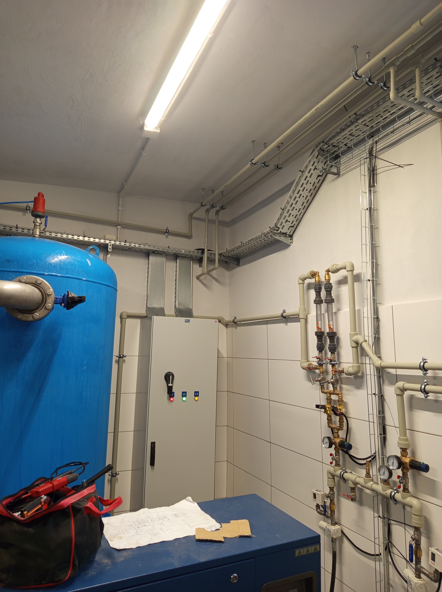 Instalacja techniczna w pomieszczeniu: niebieski zbiornik ciśnieniowy, szafa sterownicza z diodami, rury z zaworami i manometrami zamontowane na kafelkowanej ścianie, koryta kablowe pod sufitem...