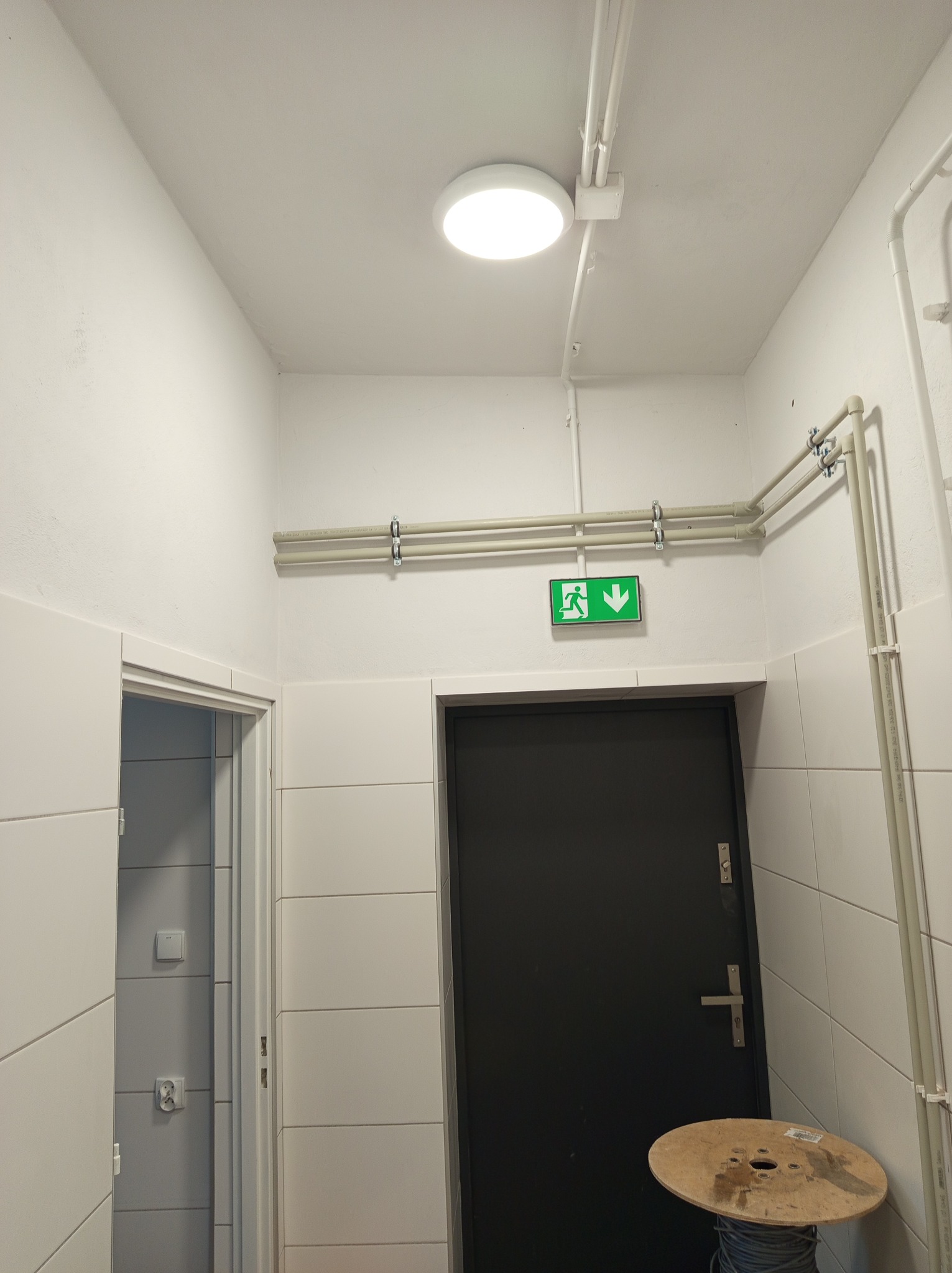 Korytarz z zamontowanym oświetleniem sufitowym, rurami instalacyjnymi przy suficie, znakiem ewakuacyjnym nad czarnymi drzwiami i szpulą kabla na podłodze.