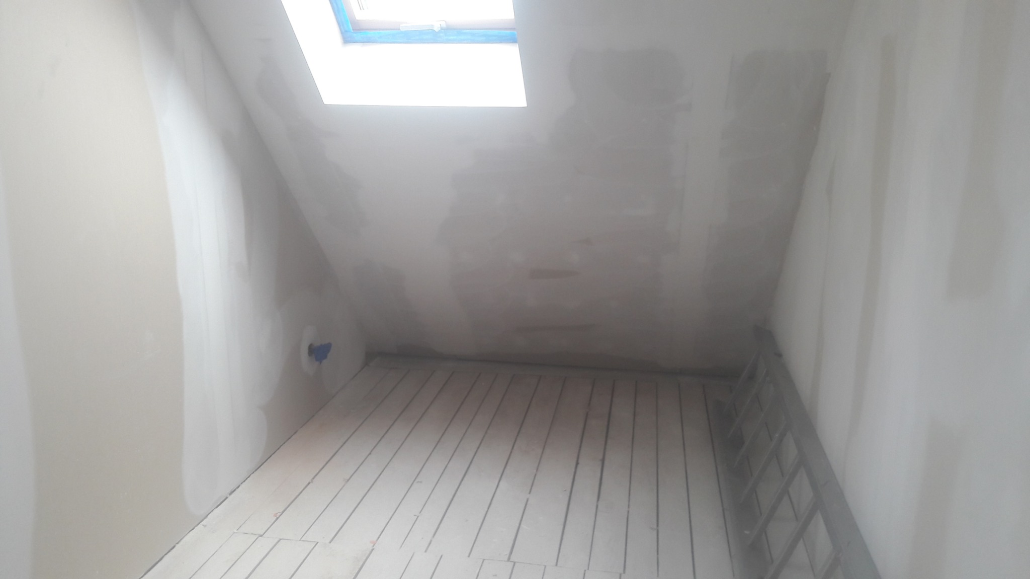 Poddasze w trakcie prac wykończeniowych: ściany pokryte gładzią gipsową, widoczne drewniane deski podłogowe i drabina oparta o ścianę, okno dachowe w górnej części kadru.