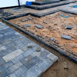 Etap prac przy układaniu szarej kostki brukowej prostokątnej na podsypce piaskowej, widoczne niezabudowane obszary, obrzeża i luźno ułożone kostki oraz niebieski przewód w piasku.