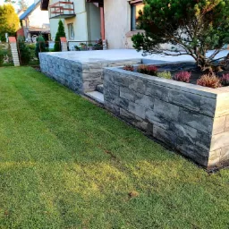 Ogród z podwyższoną rabatą z kostki brukowej, obsadzoną wrzosami i iglakiem, z równo przystrzyżonym trawnikiem w słoneczny dzień.