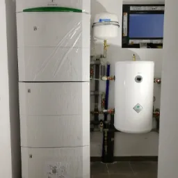 Nowa, wysoka pompa ciepła SOFATH, jeszcze w folii ochronnej, obok biały zbiornik wyrównawczy i panel sterowania w ścianie.