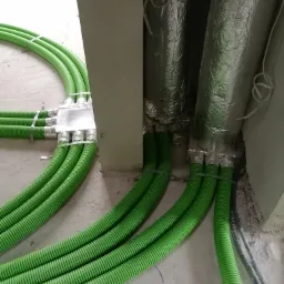 Zielone, karbowane rury wentylacyjne połączone srebrną skrzynką rozdzielczą, w tle ocieplone przewody wentylacyjne.