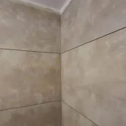 Ściana wyłożona beżowymi płytkami w dużym formacie z ciemnymi fugami, widoczny narożnik i fragment sufitu.