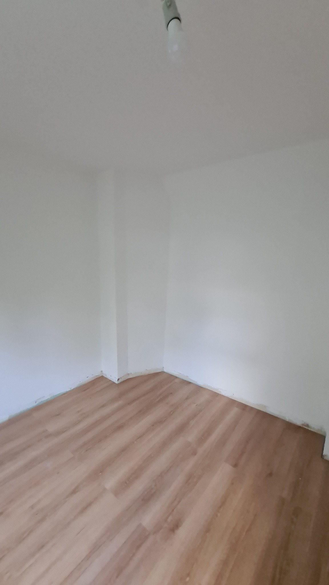 Pusty pokój po ułożeniu paneli podłogowych w odcieniach jasnego drewna, widoczne białe ściany po świeżym malowaniu i surowa żarówka na suficie.