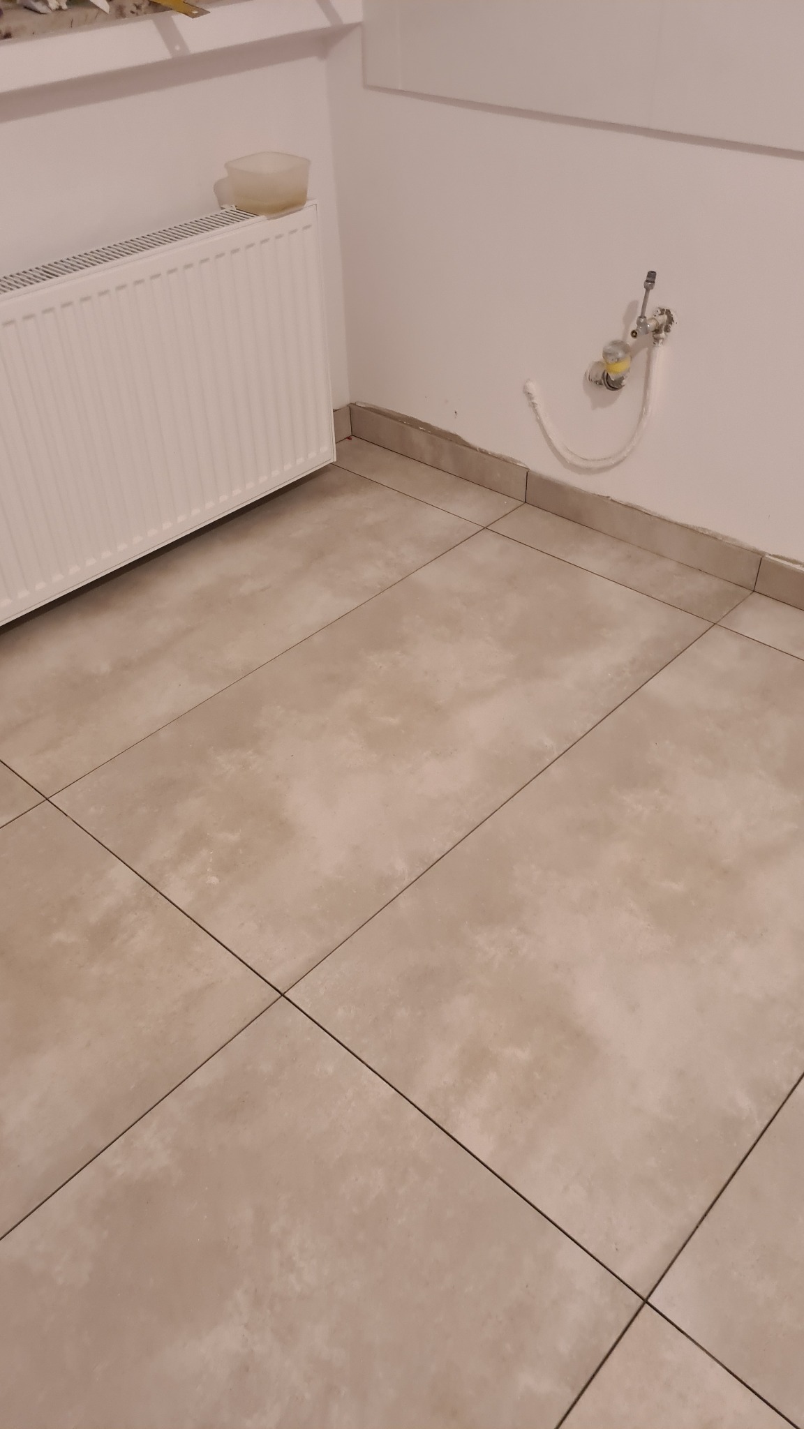 Ułożone, beżowe płytki podłogowe w łazience z grzejnikiem i rurami doprowadzającymi wodę wystającymi ze ściany.