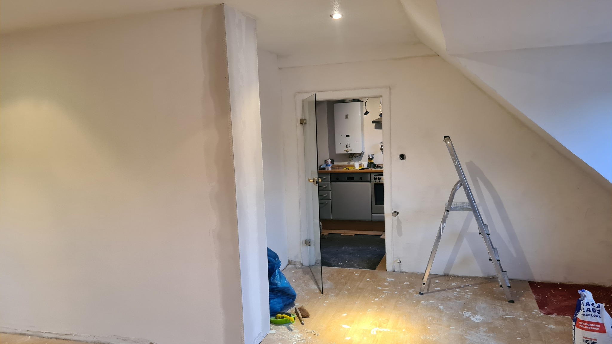 Pokój na poddaszu w trakcie prac: ściany po szpachlowaniu, widoczna drabina, przez otwarte drzwi widoczny fragment kuchni z piecem gazowym.