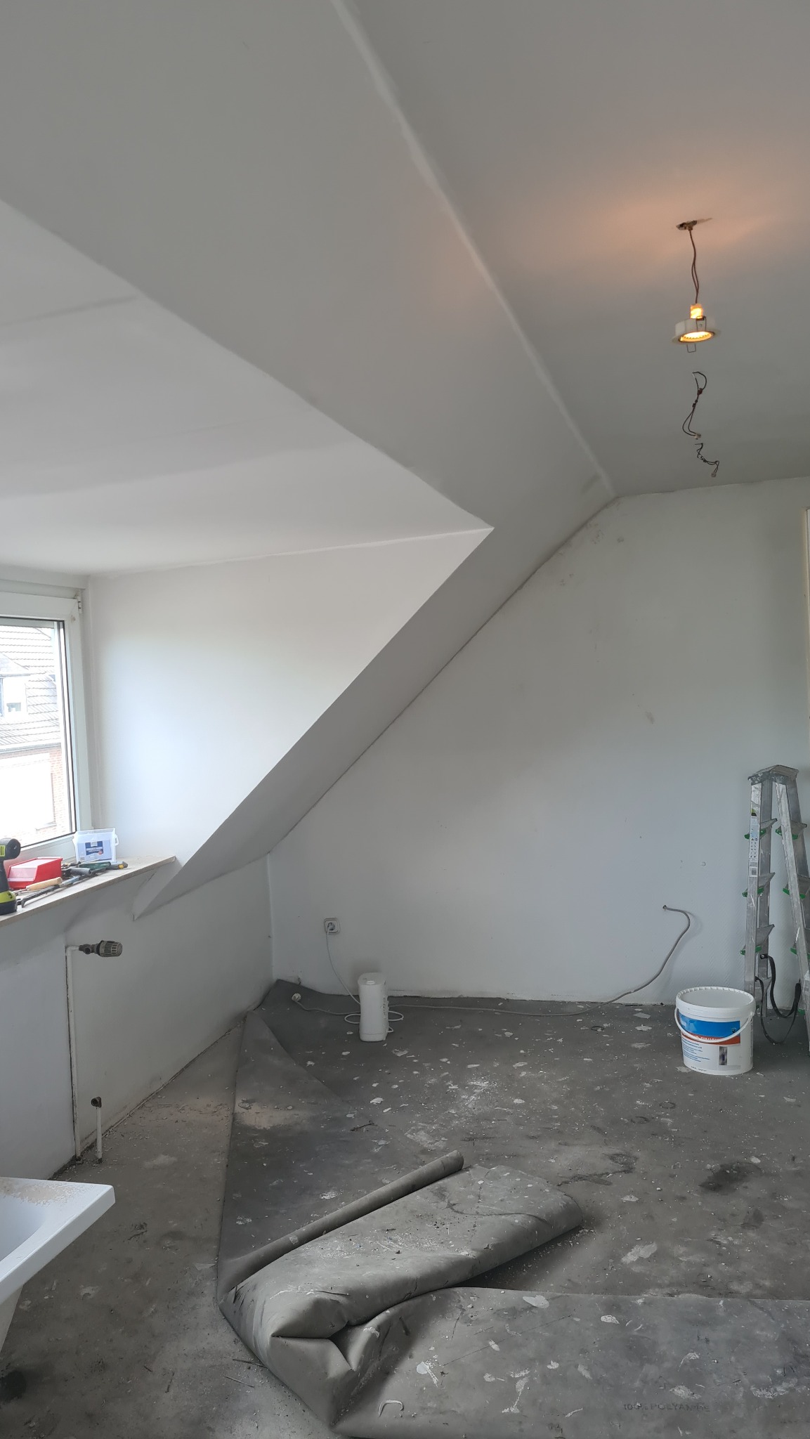 Pokój na poddaszu w trakcie prac, z widoczną białą ścianą, oknem, grzejnikiem, zabezpieczoną podłogą, wiadrem z farbą i rozłożoną drabiną.