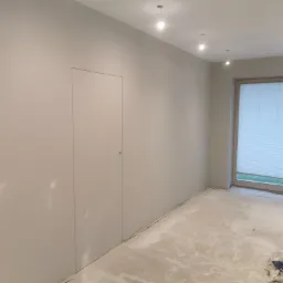 Pomalowane na szaro wnętrze z ukrytymi drzwiami i oknami z żaluzjami, podłoga przygotowana do dalszych prac wykończeniowych.