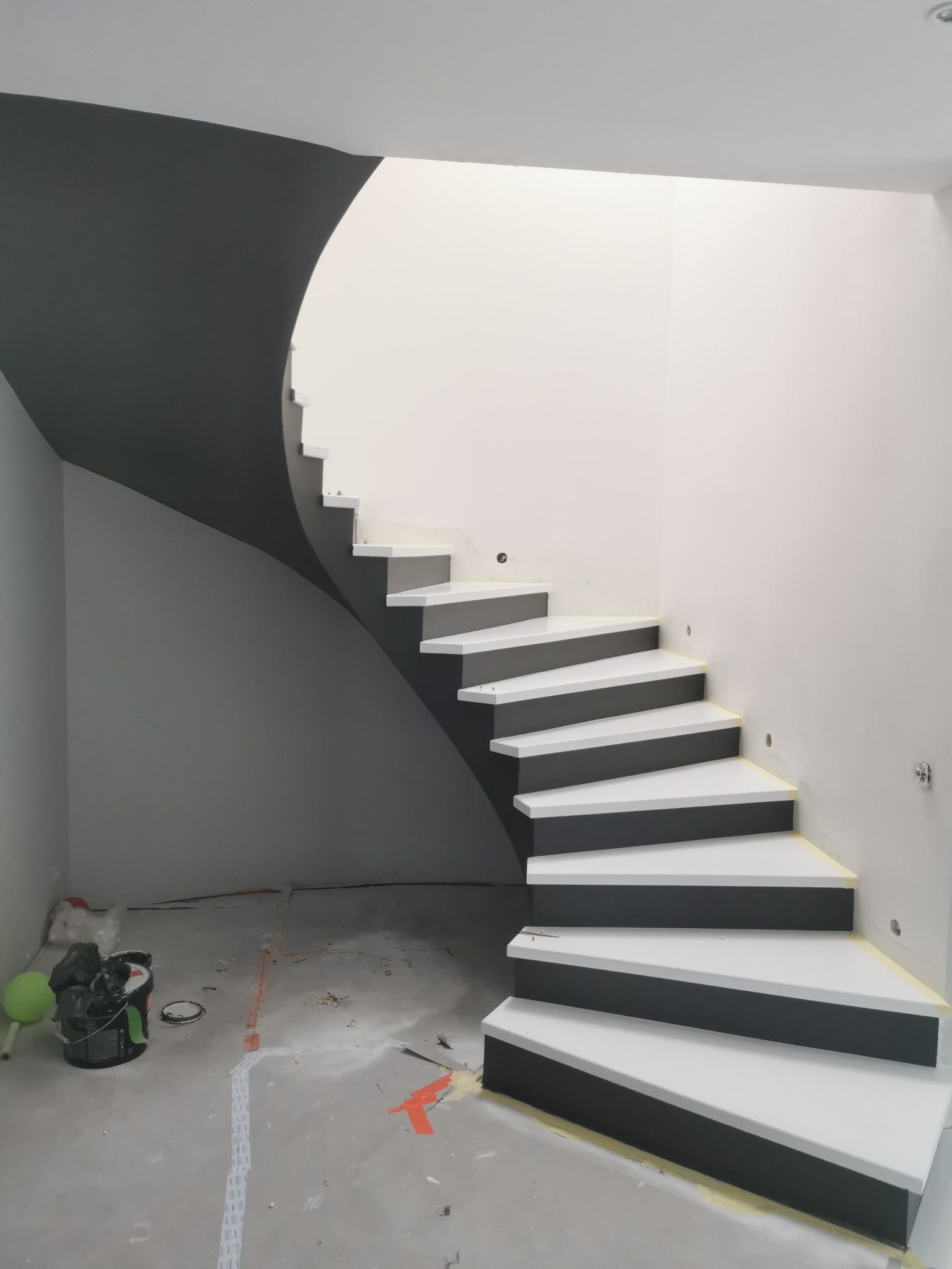 Nowoczesne, spiralne schody z białymi stopniami i ciemnoszarymi bokami, kontrastujące z białą ścianą i fragmentem ciemnoszarej ściany. Widoczne ślady prac wykończeniowych na podłodze.