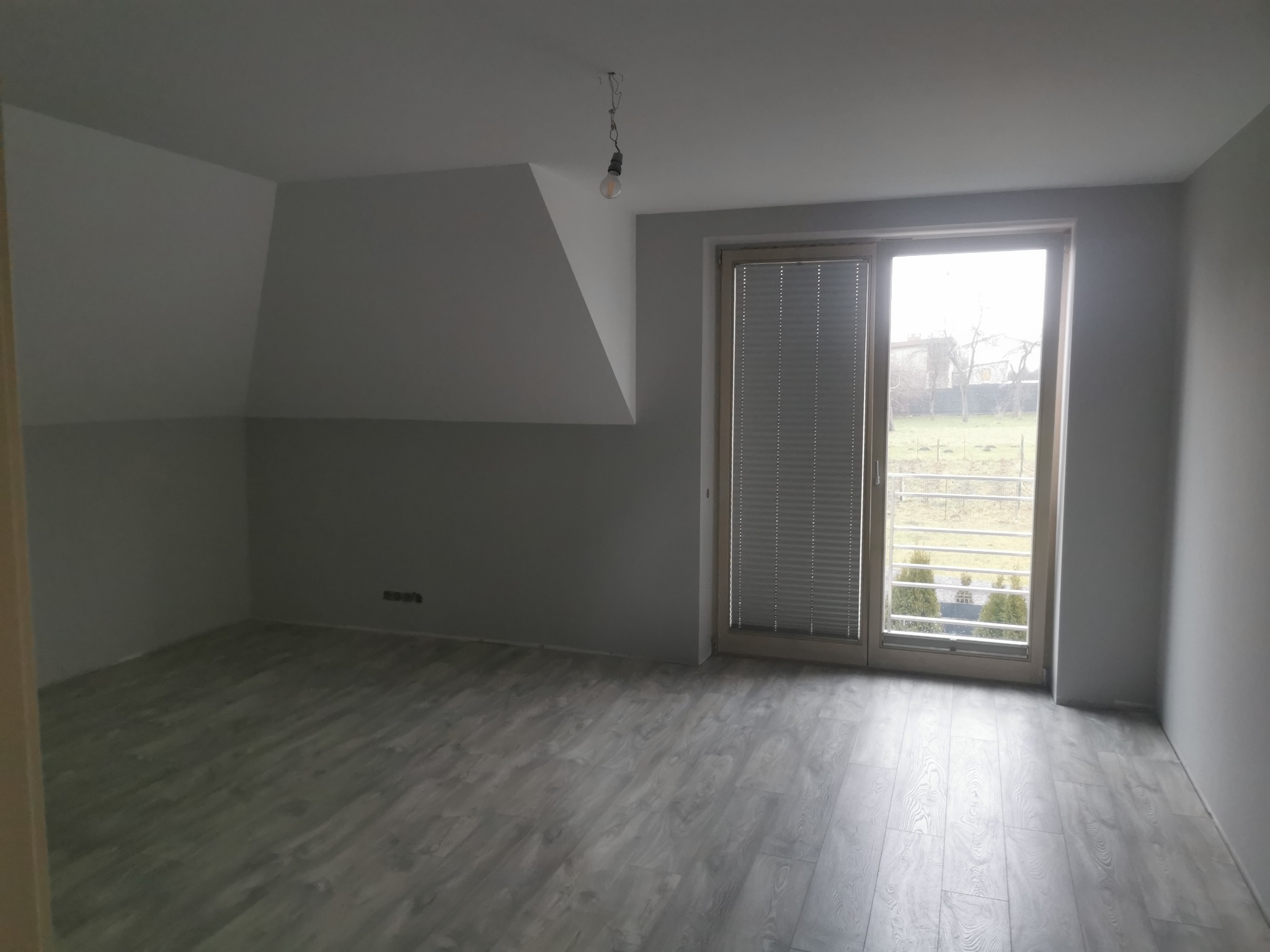 Pusty pokój na poddaszu z szarymi ścianami, drewnopodobną podłogą, oknem z roletami i wiszącą żarówką bez klosza.