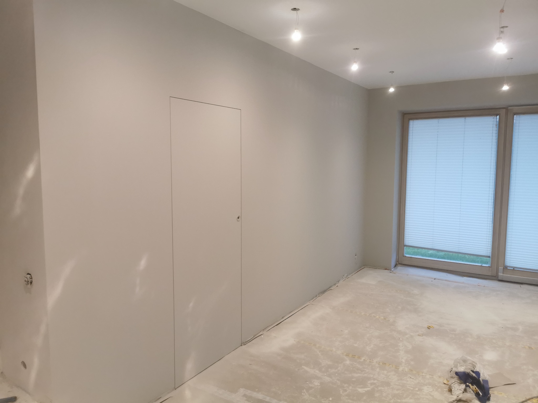 Pomalowane na szaro wnętrze z ukrytymi drzwiami i oknami z żaluzjami, podłoga przygotowana do dalszych prac wykończeniowych.