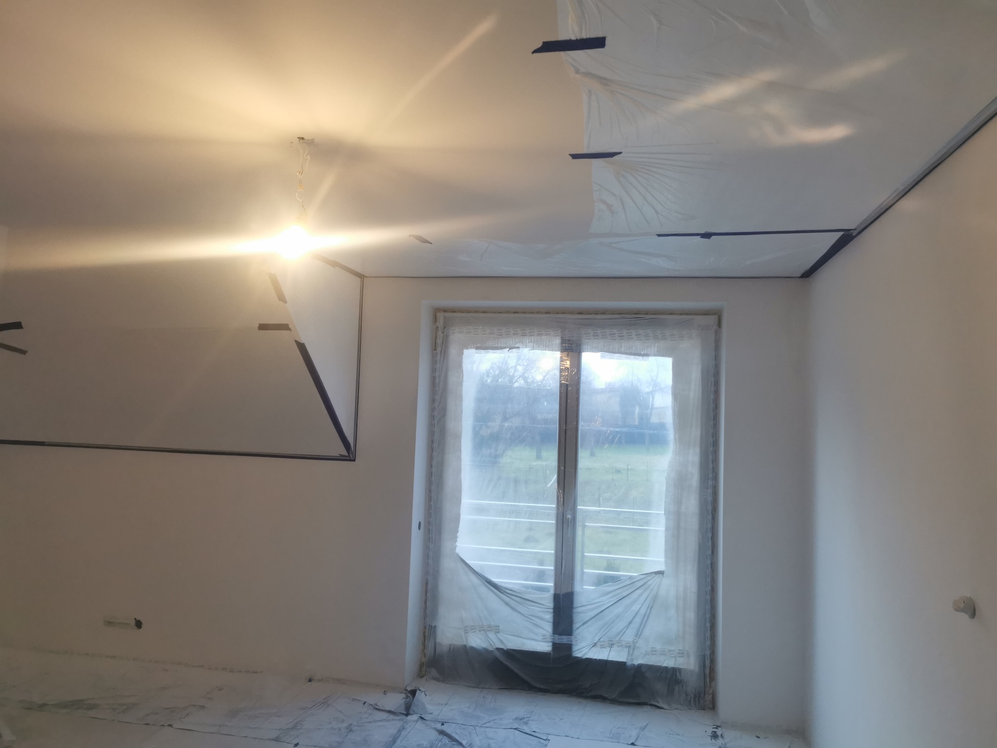 Pokój przygotowany do malowania: podłoga i okna zabezpieczone folią, ściany i sufit świeżo zagruntowane, widoczne taśmy malarskie.