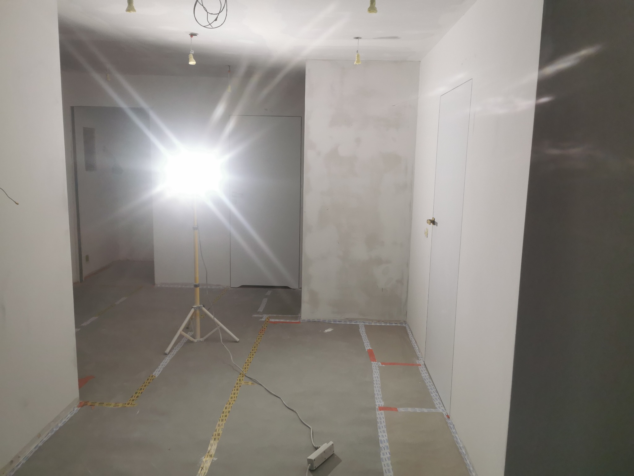 Przygotowane do prac wykończeniowych pomieszczenie z surowymi ścianami, podłoga z wyznaczonymi strefami taśmą, oświetlane lampą budowlaną na statywie.