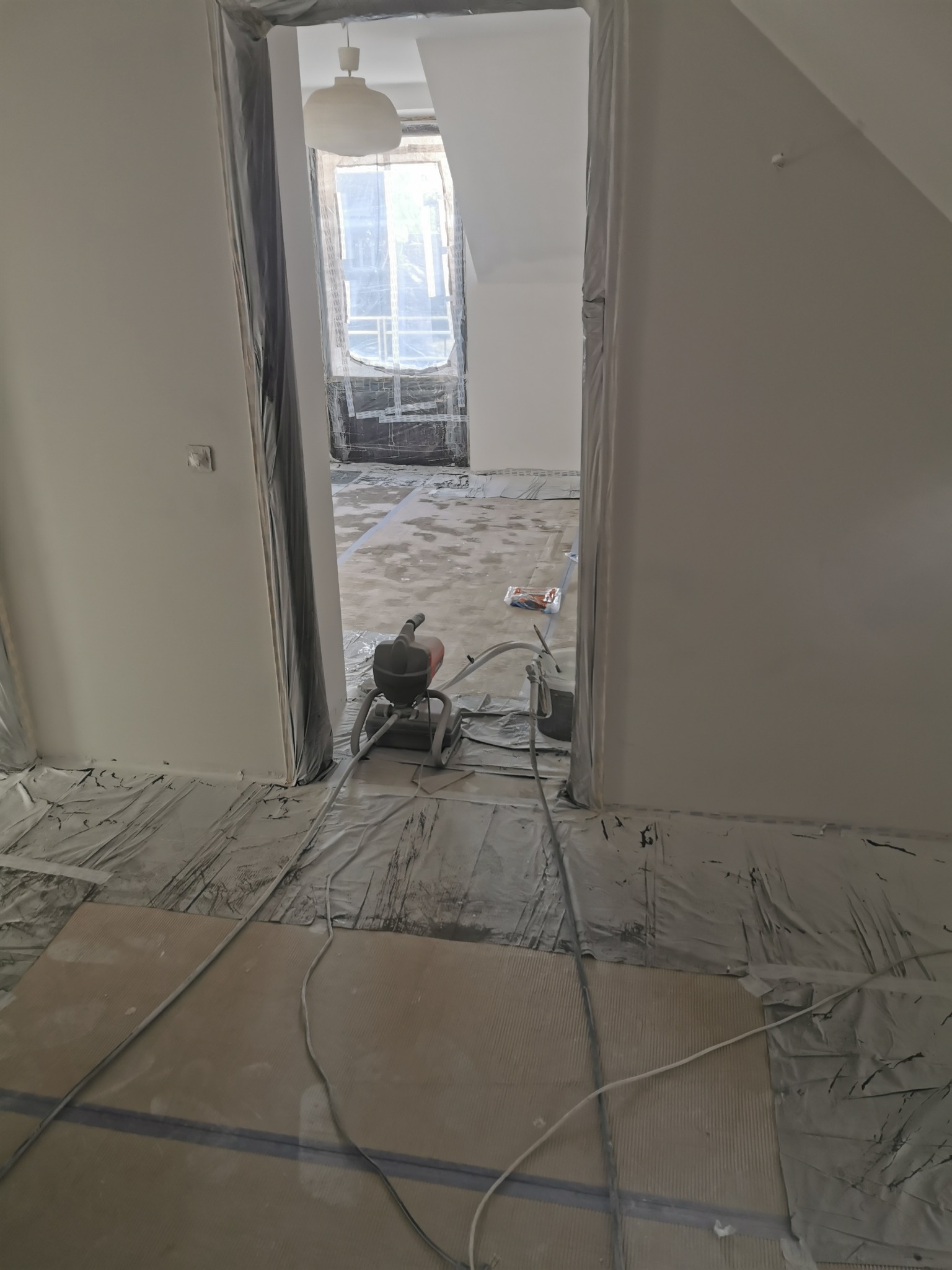 Wnętrze w trakcie prac przygotowawczych do malowania, z maszyną natryskową na podłodze zabezpieczonej folią, widoczne osłonięte folią drzwi i okno.