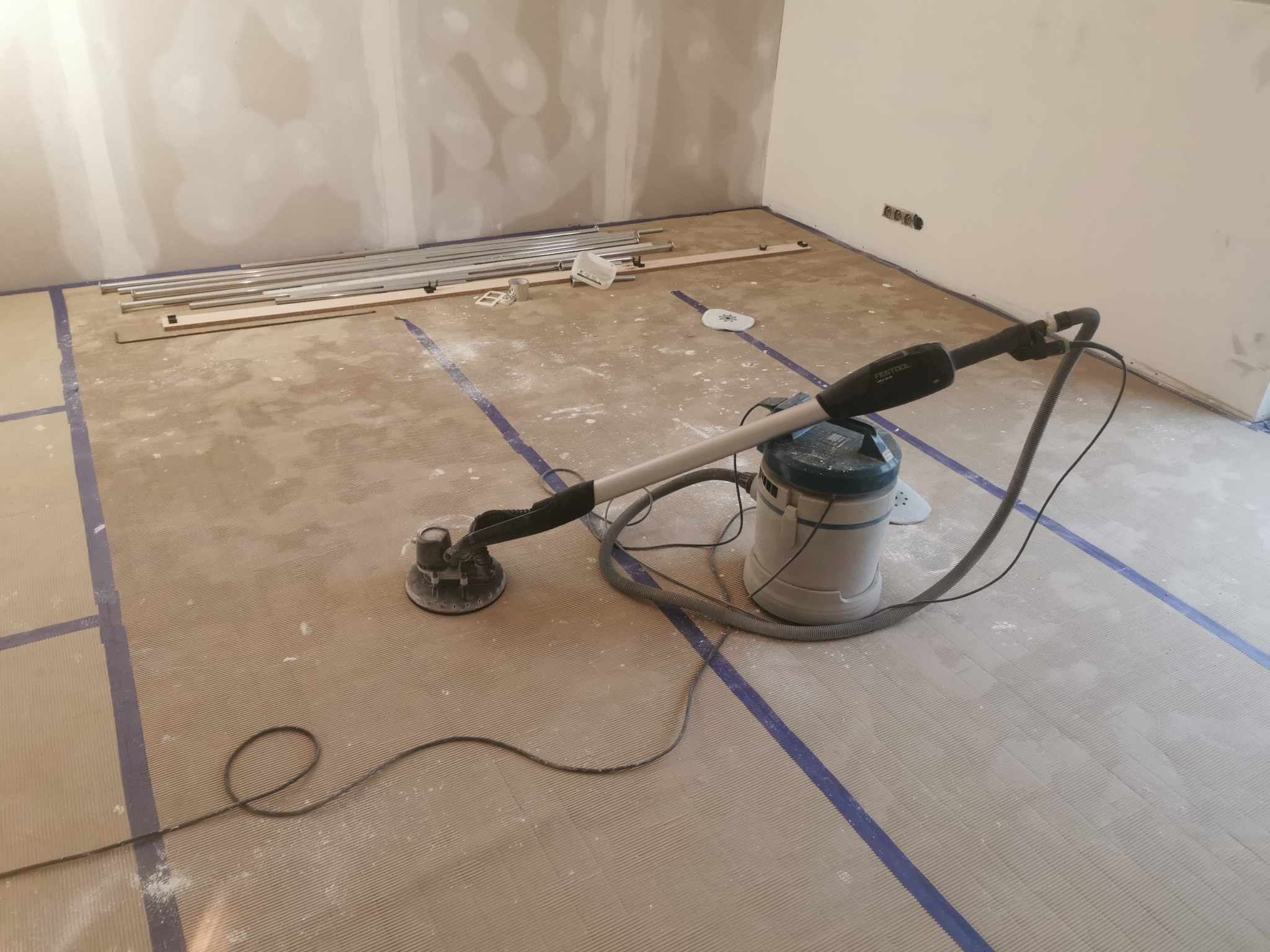 Szlifierka oscylacyjna marki Festool na podłodze z wylewką samopoziomującą, zabezpieczonej niebieską taśmą, w trakcie prac wykończeniowych w pomieszczeniu z płytami kartonowo-gipsowymi na ścianach.