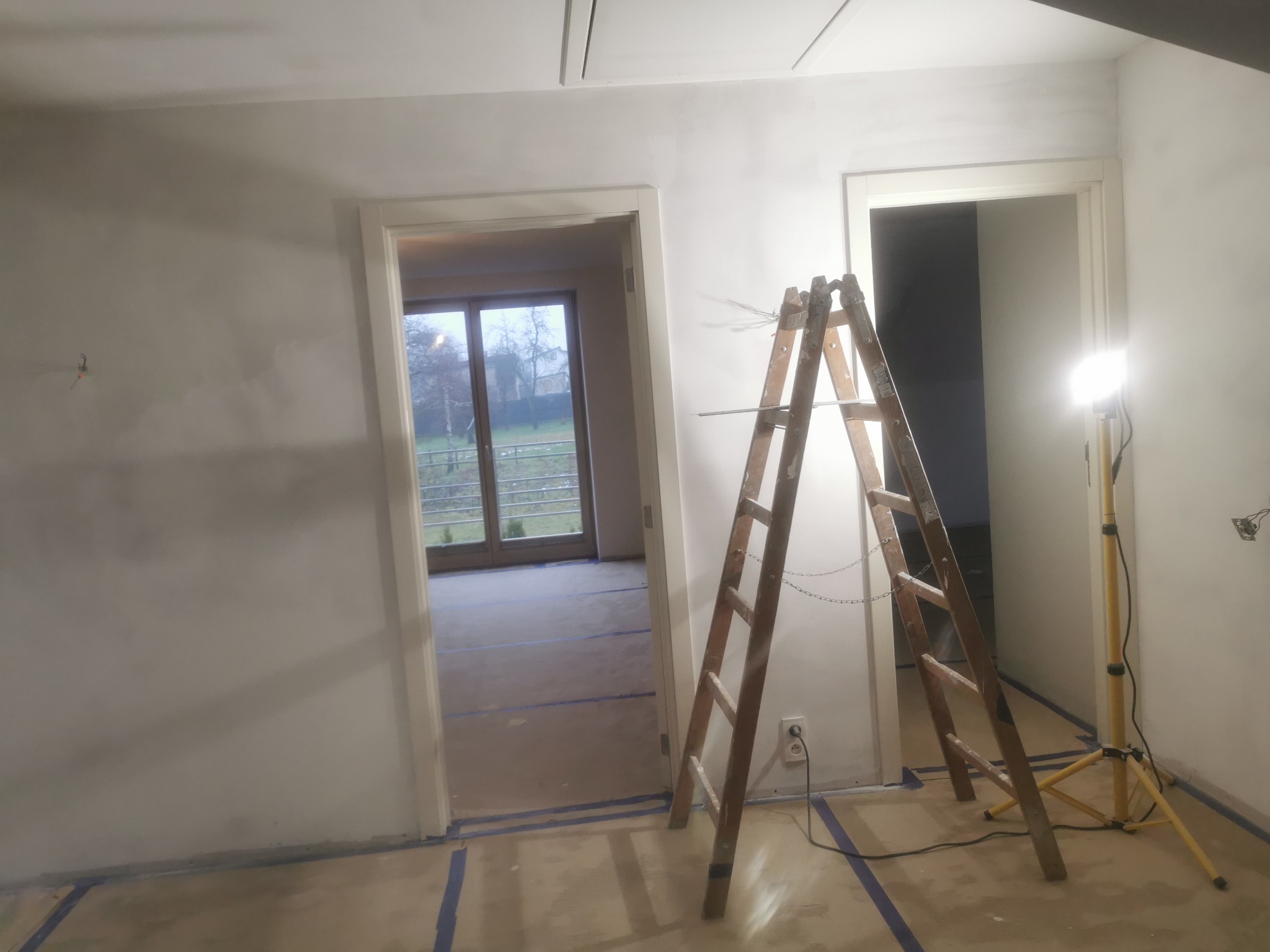 Pokój w trakcie odświeżania: świeżo wyszpachlowane ściany, podłoga zabezpieczona taśmą malarską, drabina oparta o ścianę i halogen na statywie oświetlający wnętrze.