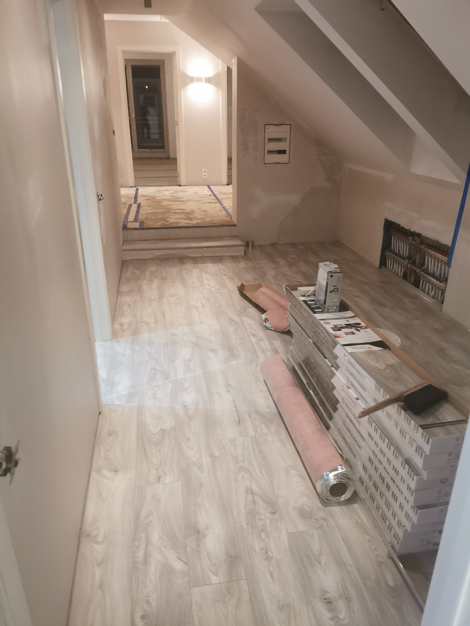 Poddasze w trakcie prac wykończeniowych: ułożona podłoga z paneli, przygotowane paczki paneli, widoczna instalacja grzewcza w ścianie, ściany przygotowane do malowania, widoczna skrzynka elektryczna.