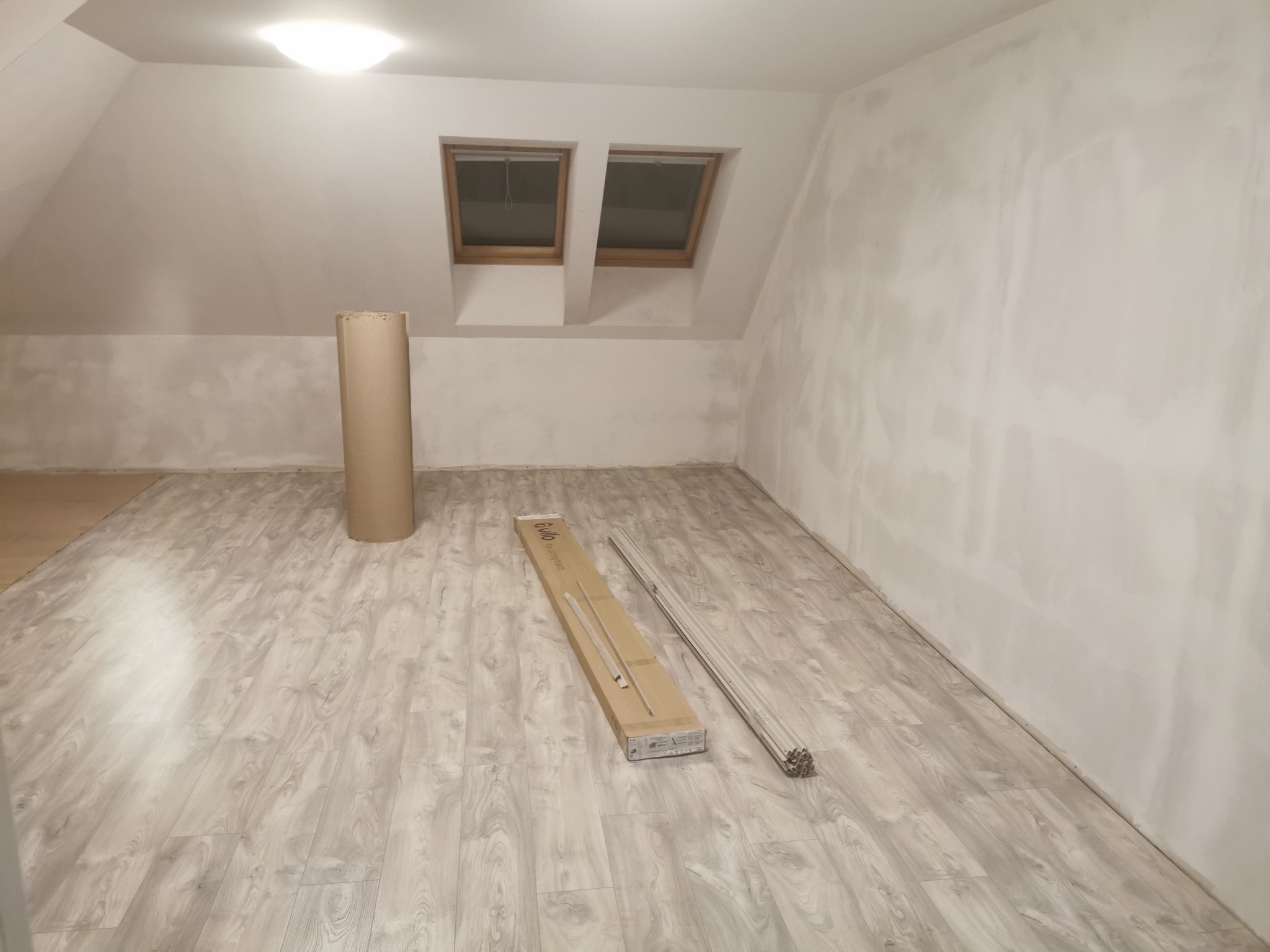 Poddasze w trakcie przygotowania do prac wykończeniowych: zagruntowane ściany, panele podłogowe w odcieniach szarości, paczka z listwami przypodłogowymi firmy Evilo i rura z tektury. Dwa okna...