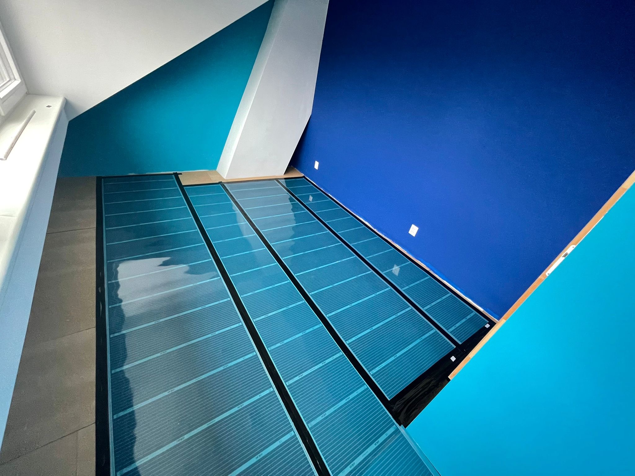 Nietypowe ujęcie z góry na zainstalowane, niebieskie maty grzewcze na podłodze, z widocznymi fragmentami ścian w odcieniach błękitu i bieli, oraz szarą podłogą w tle.