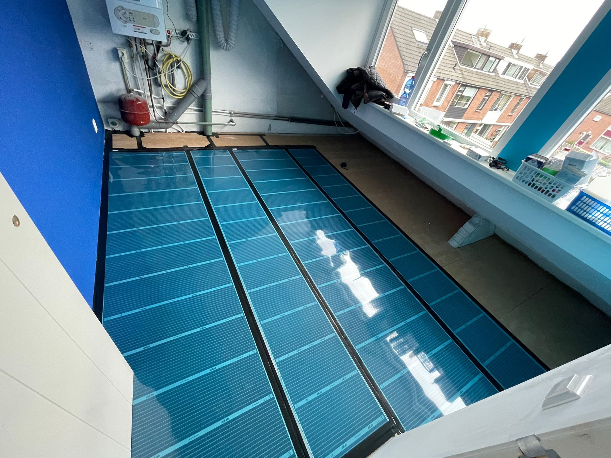 Błyszczące, niebieskie maty grzewcze rozłożone na drewnianej podłodze pod skosem, widok z góry, instalacja w trakcie, widoczne elementy instalacji CO na ścianie.