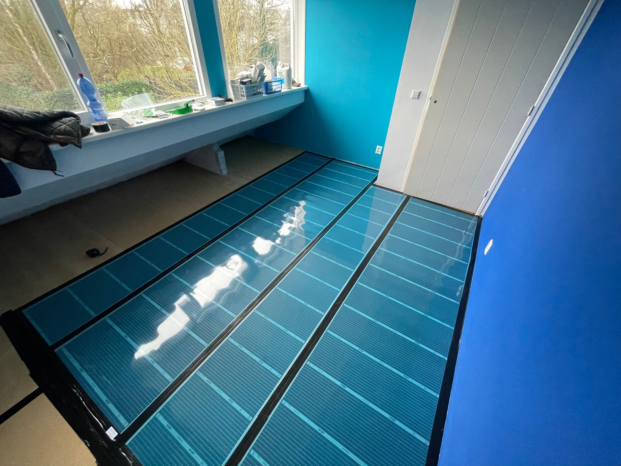 Niebieskie maty grzewcze rozłożone na podłodze w pokoju z oknem i niebieskimi ścianami, przygotowane do montażu paneli podłogowych.